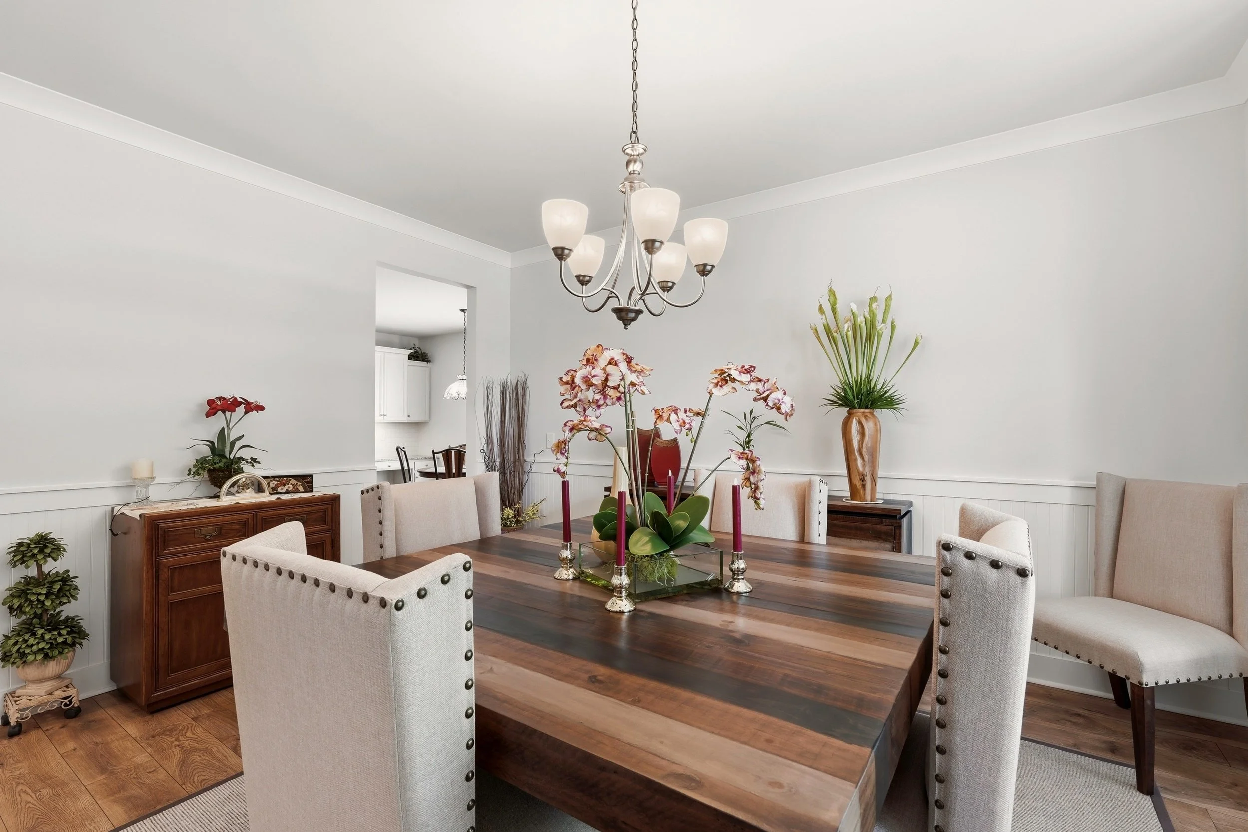 Dining room with a large wooden table surrounded by beige upholstered chairs, floral centerpieces, and potted plants on side tables and a cabinet, lit by a chandelier hanging from the ceiling.