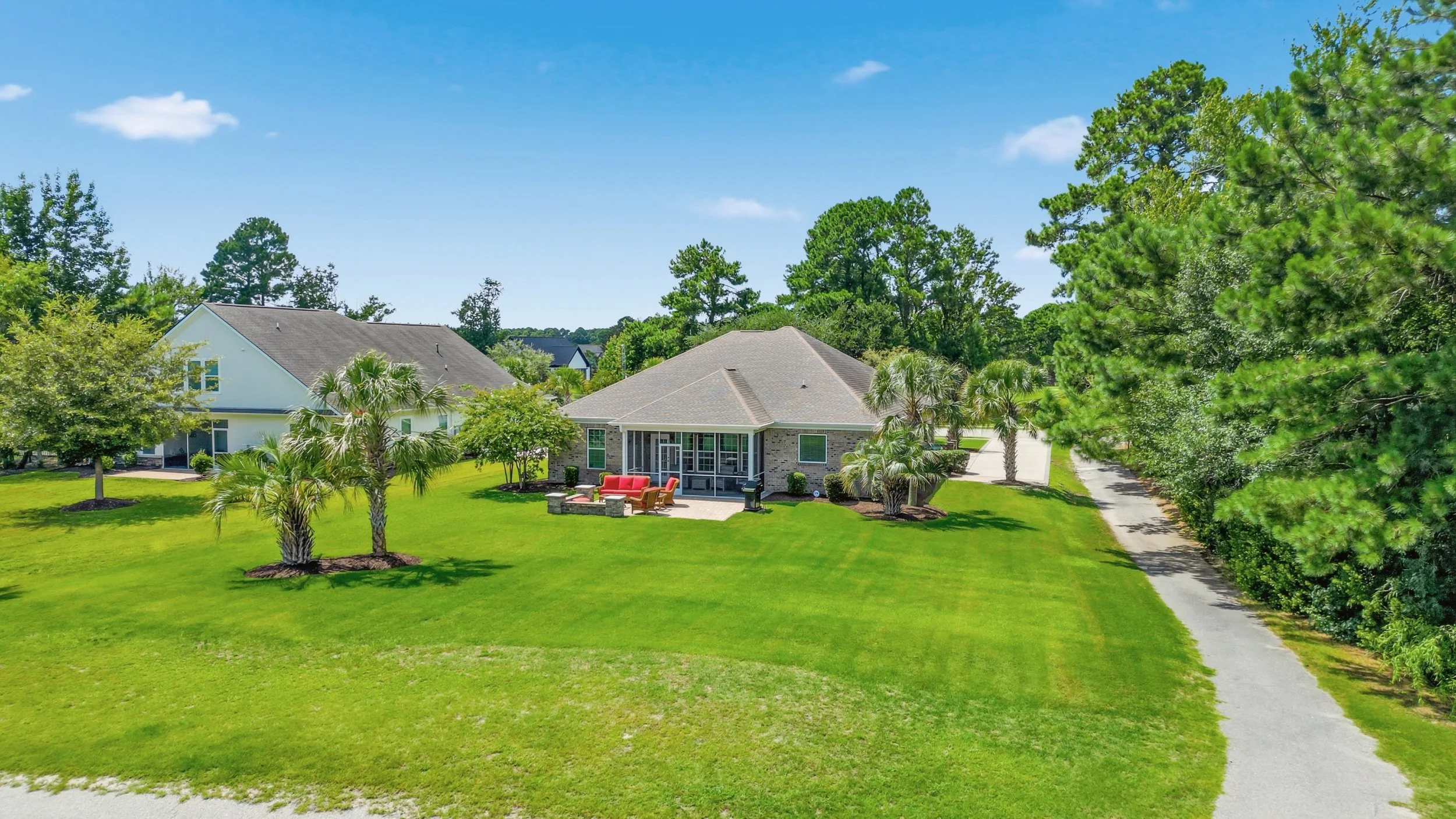 Lush green lawn with a brick house in the background, surrounded by tall trees and a paved driveway on the right side.