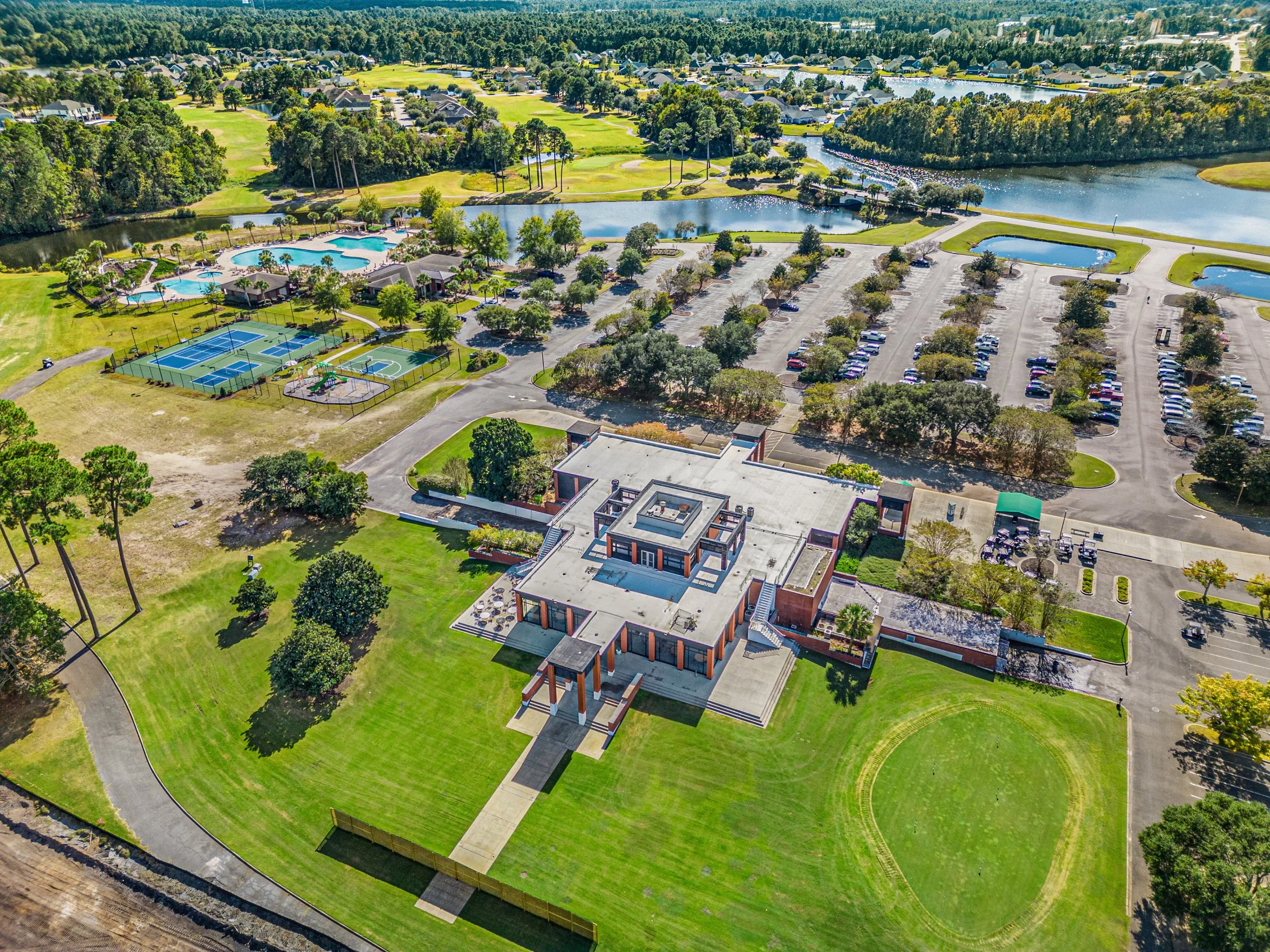 An aerial view of a residential community featuring a large park with multiple swimming pools, tennis courts, a pond, and parking lots, surrounded by houses and trees.