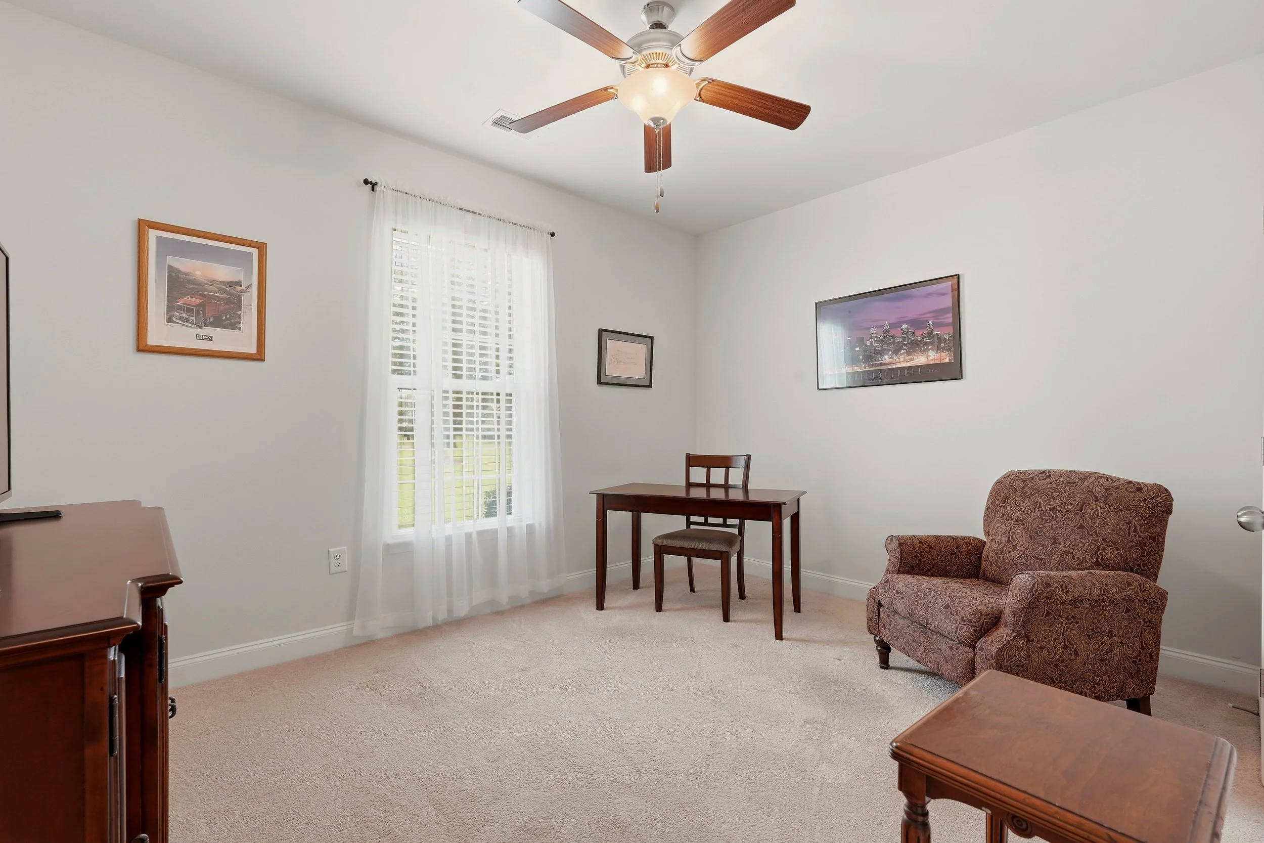 Living room with a ceiling fan, framed pictures on white walls, a window with sheer curtains, and furniture including a patterned armchair and wooden tables.