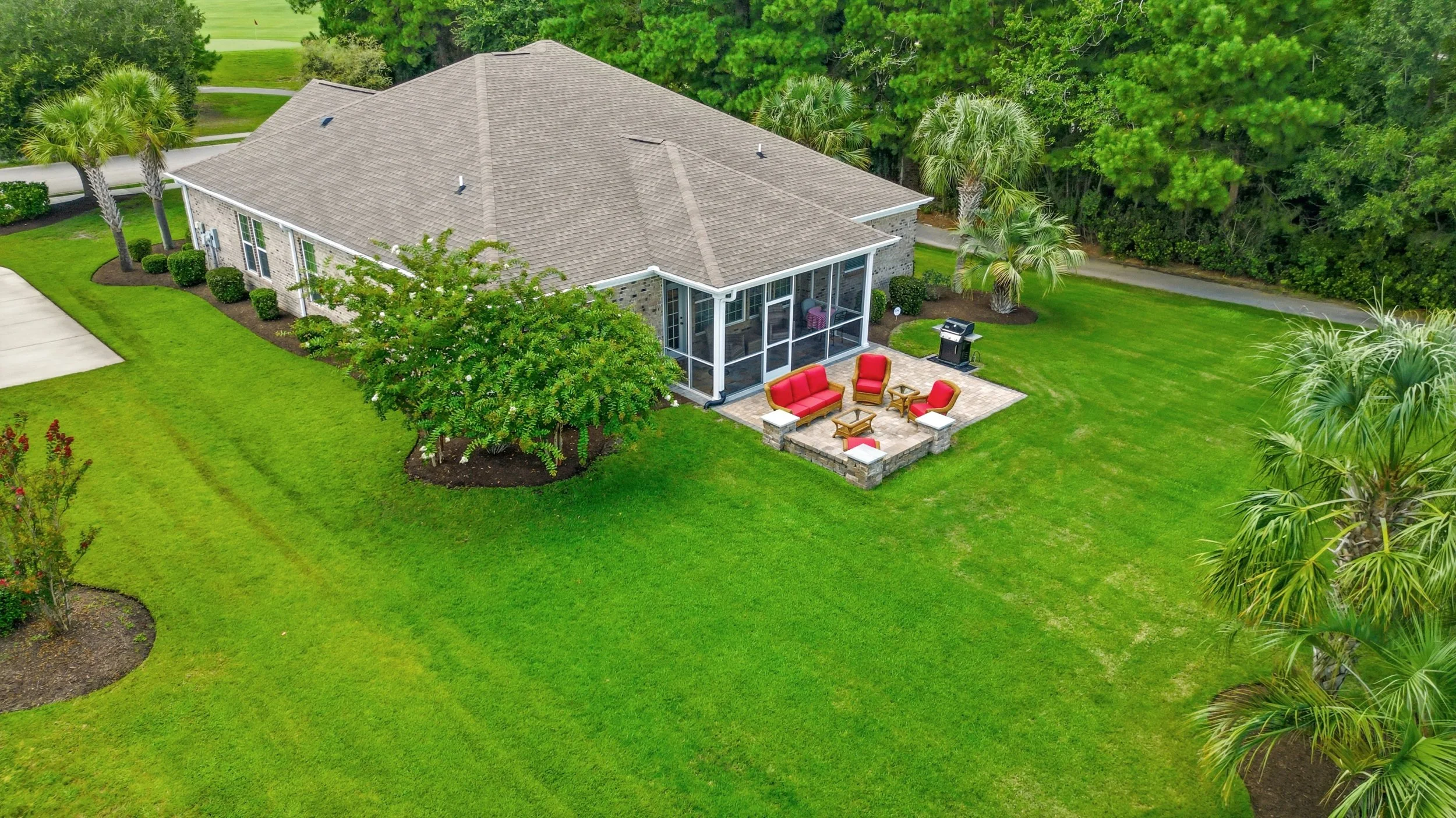 Aerial view of a house with a screened porch, outdoor seating with red cushions, and a grassy backyard with trees and landscaped garden beds.