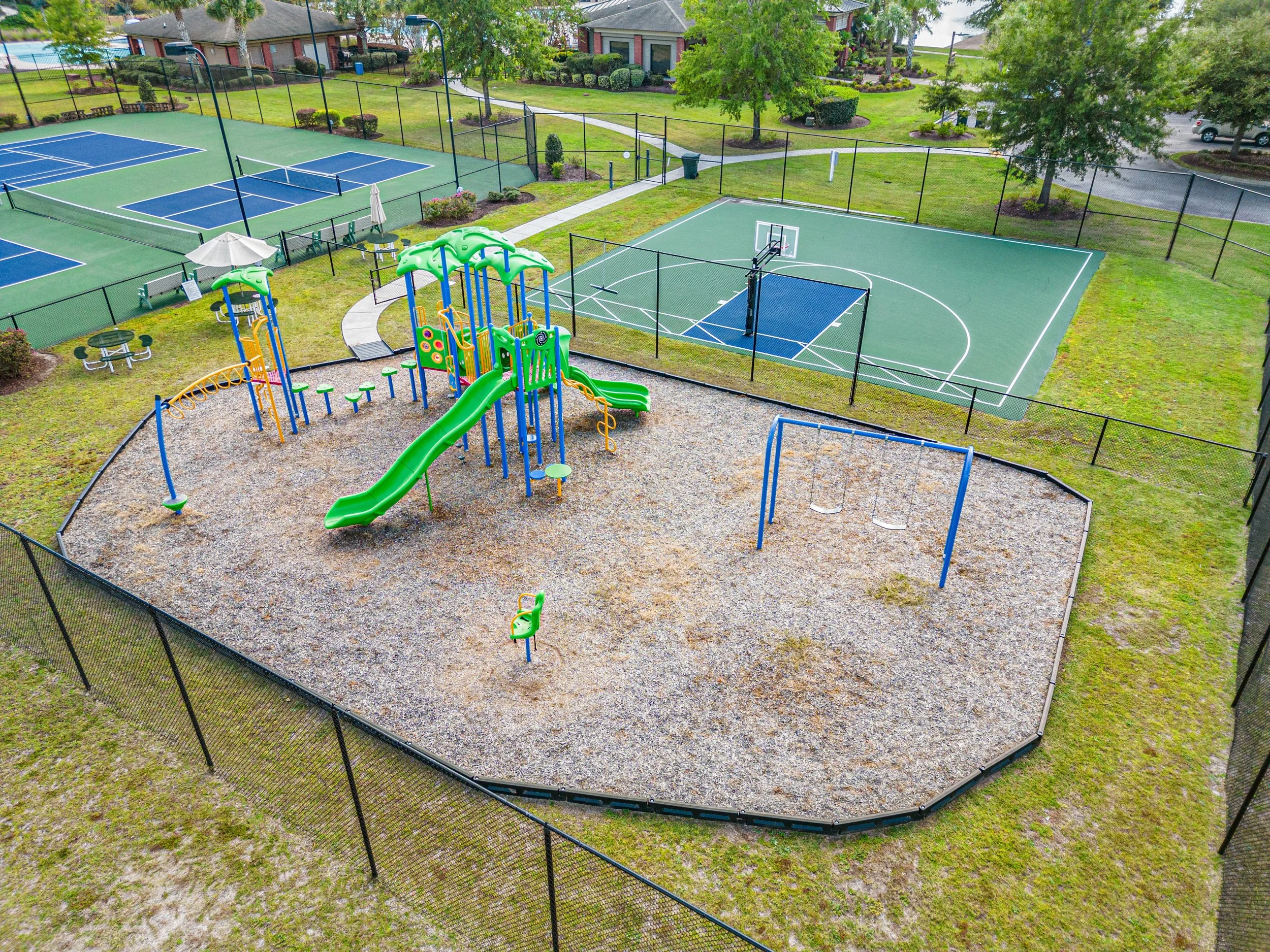 An outdoor sports and playground area within a residential complex, featuring multiple tennis courts, a playground with slides and climbing structures, picnic tables, surrounded by green grass, trees, and residential buildings in the background.