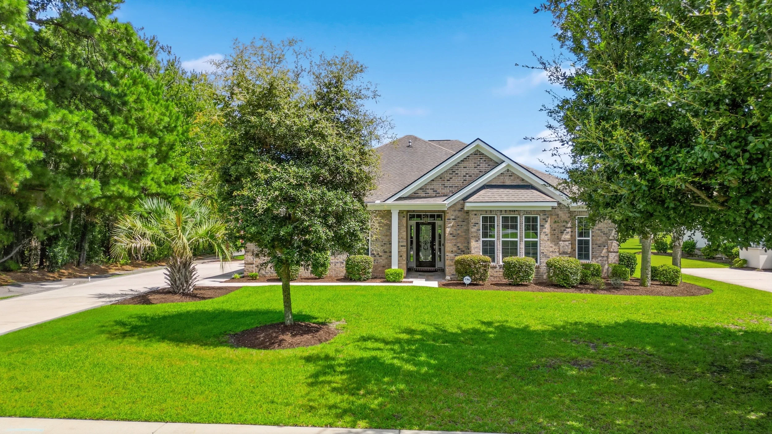 A suburban house with a brick exterior and a gabled roof, surrounded by a well-maintained lawn and various trees including a pine and a palm tree. The house has large windows and a front porch with a black door.
