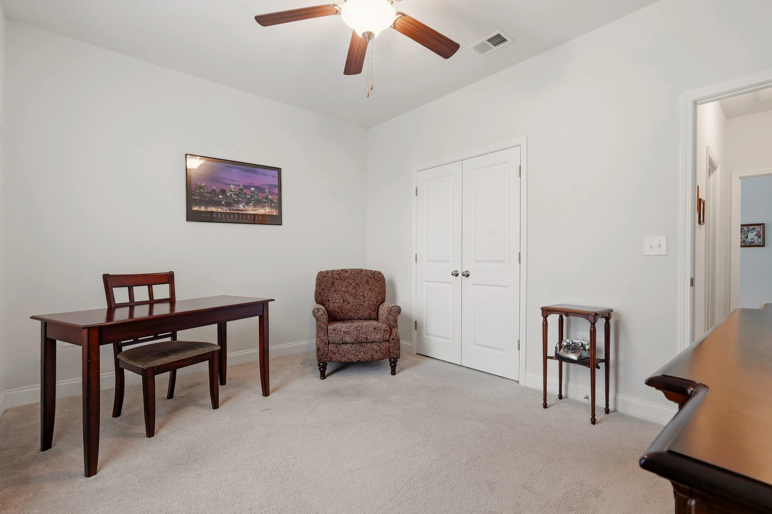 A room with white walls, a ceiling fan with wooden blades, framed cityscape art on the wall, a wooden dining table with chairs, an upholstered armchair, and a small side table with a vintage rotary phone. There are closed white closet doors and an el