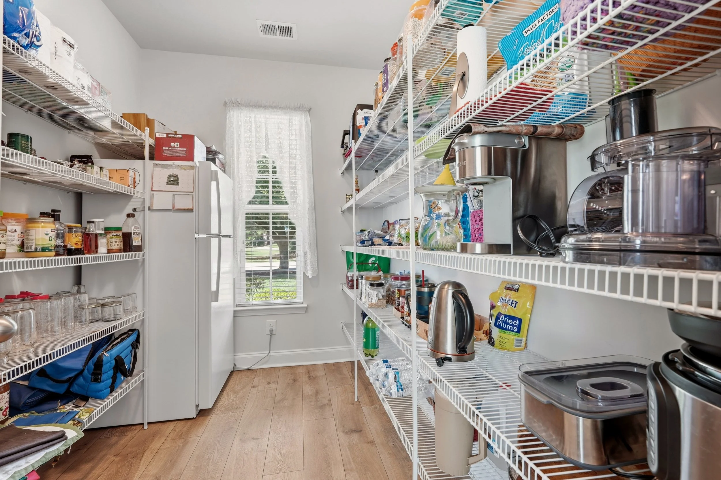A small pantry with white wire shelves holding food items, kitchen appliances, and glassware. There are cabinets, a window, and a fridge in the background.