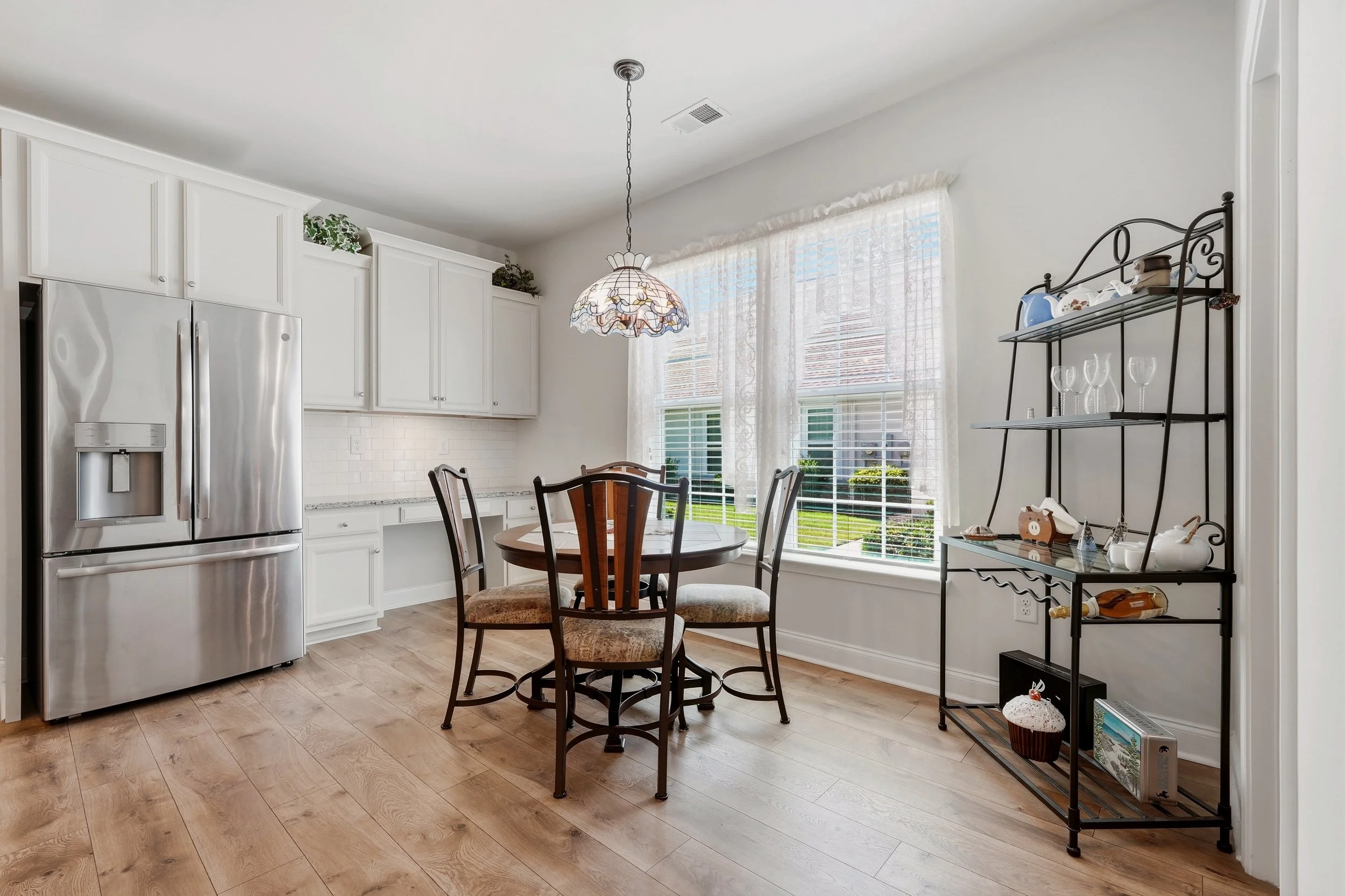 Bright kitchen with white cabinets, stainless steel refrigerator, wooden dining table with four chairs, large window with sheer curtains, and a black metal shelf with glassware and decorative items.