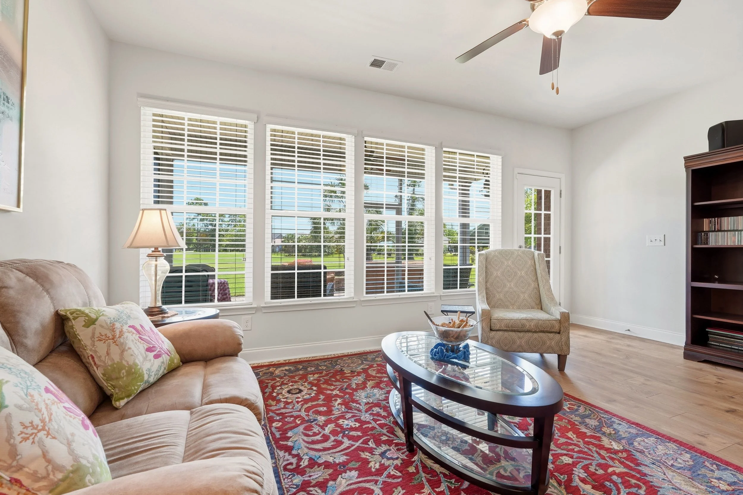 Living room with large windows, beige sofa with floral pillows, glass coffee table, upholstered armchair, wooden bookshelf, ceiling fan, and a view of a green yard outside.