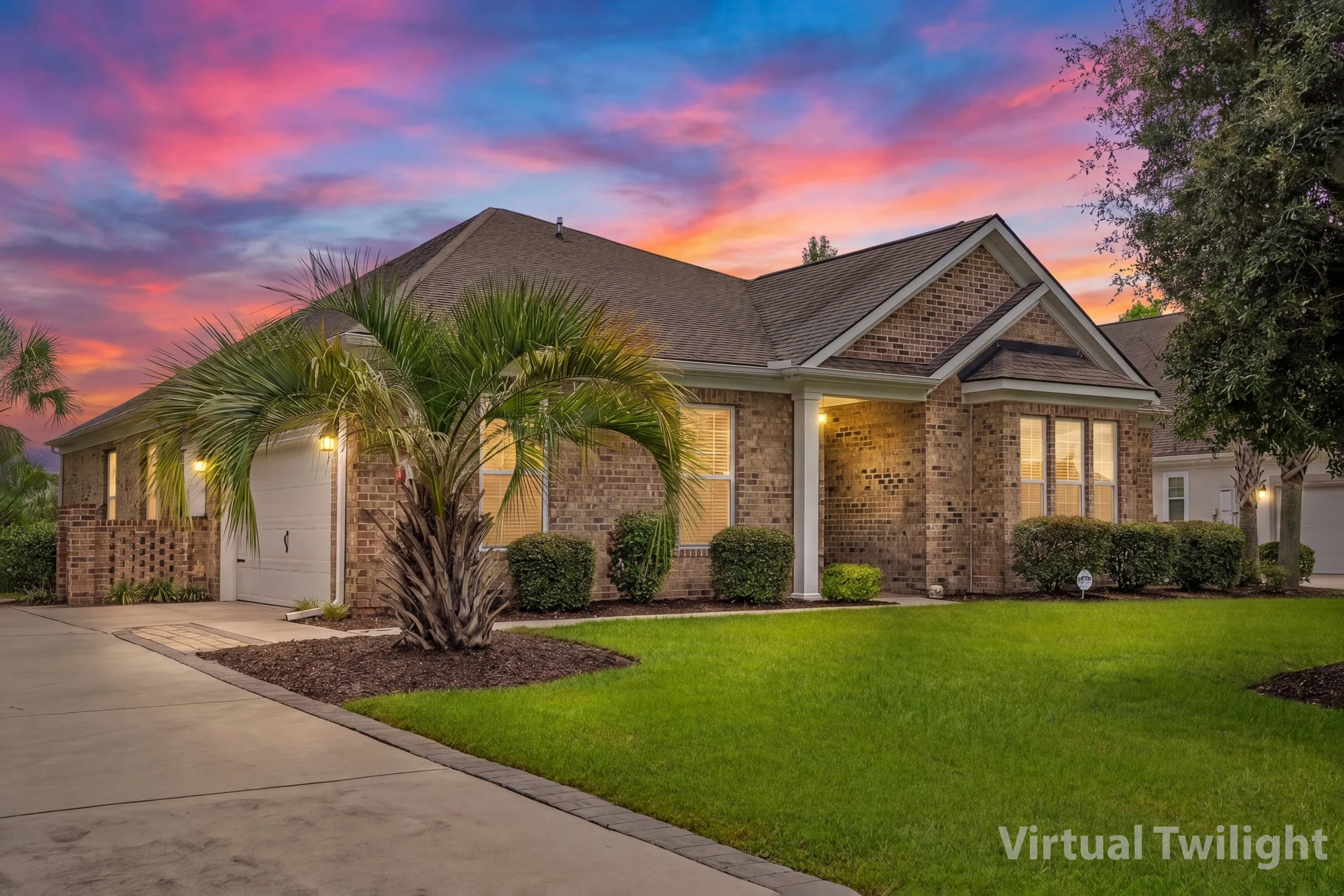 A brick house with a peaked roof at sunset, with trees and shrubs in the front yard, and lights on inside.