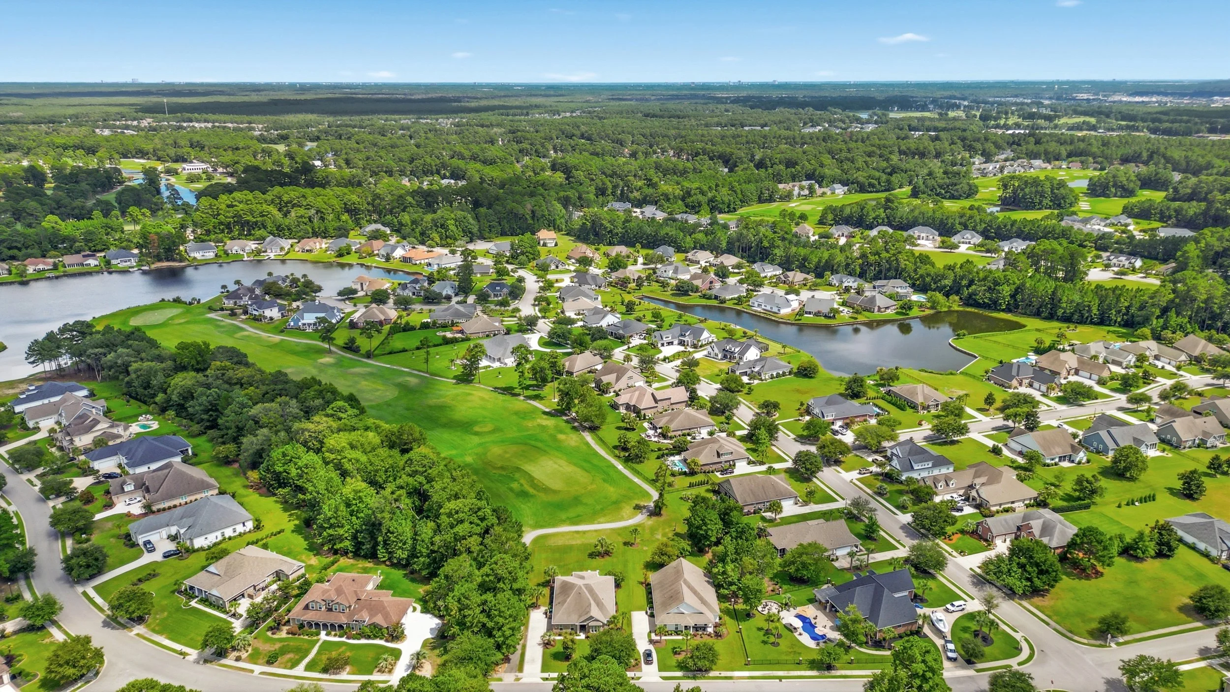 Aerial view of a suburban neighborhood with lakes, green lawns, trees, and houses, surrounded by forested areas.