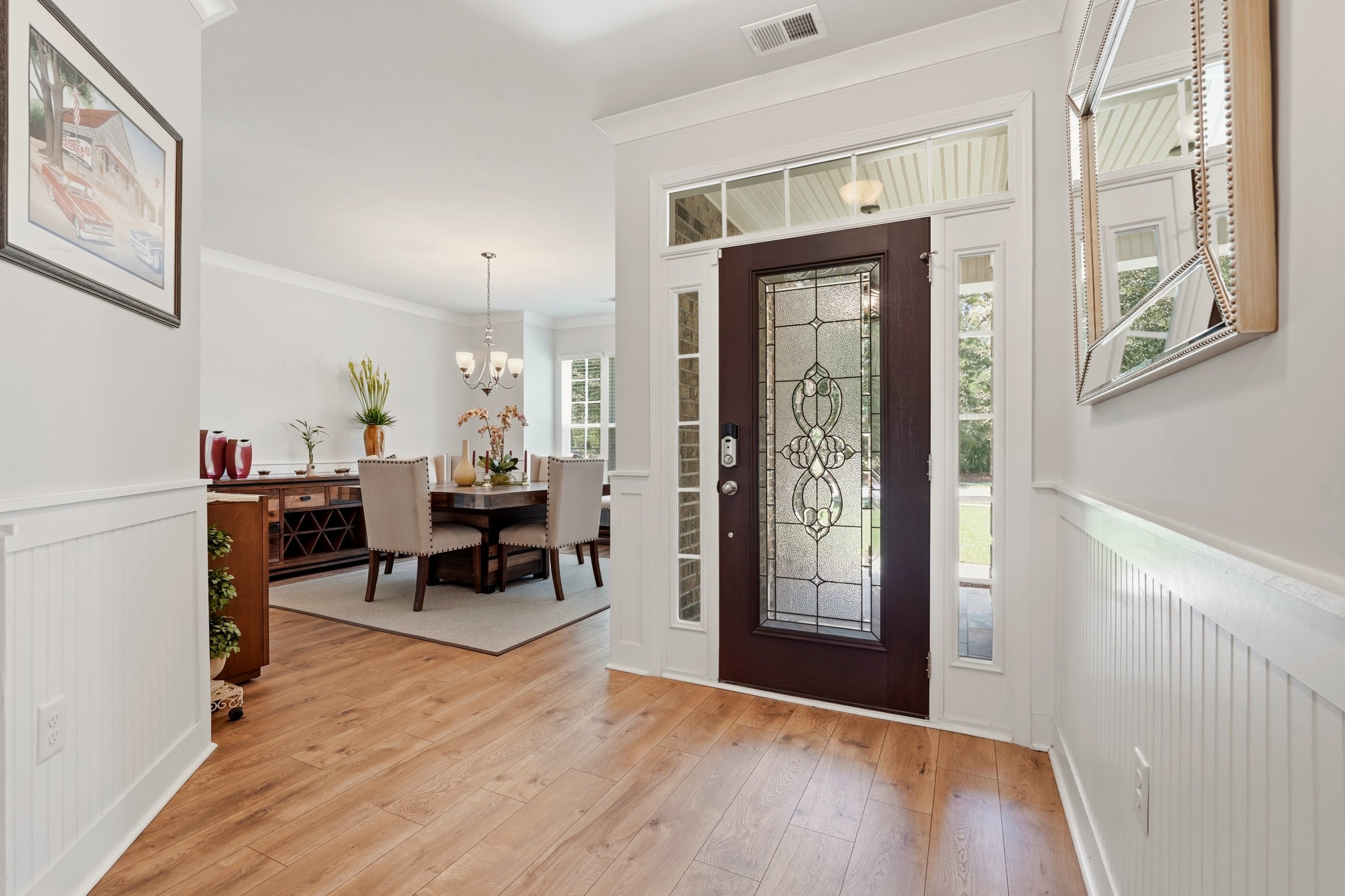 Interior of a house entryway with a front door that has decorative glass, side windows, and a transom window. To the left, a dining area with a wooden table, beige chairs, a chandelier, and a sideboard with decorative vases and plants. Hardwood floor