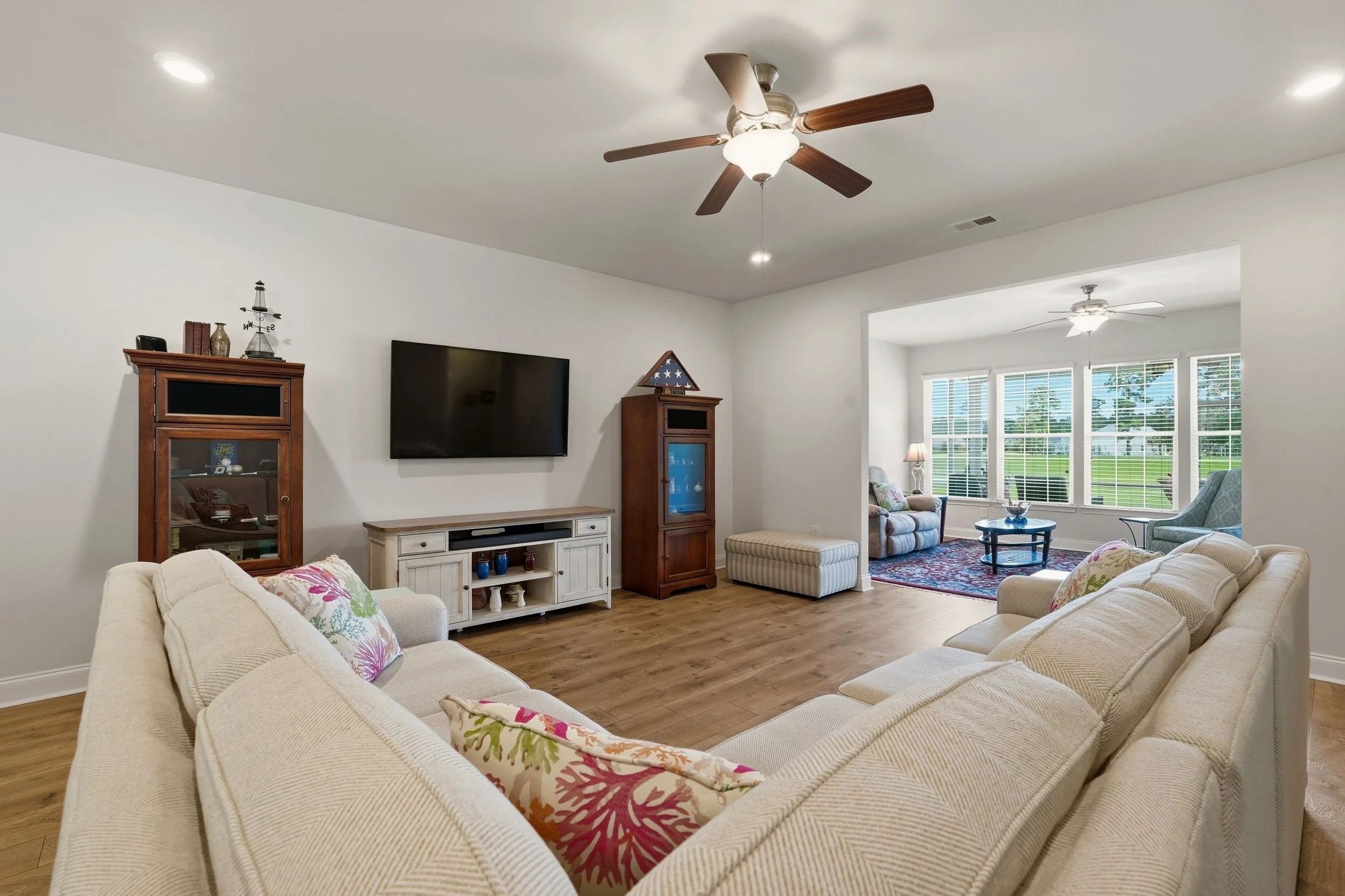 Living room with beige sofas, floral pillows, wooden cabinets, a wall-mounted TV, and large windows with blinds.