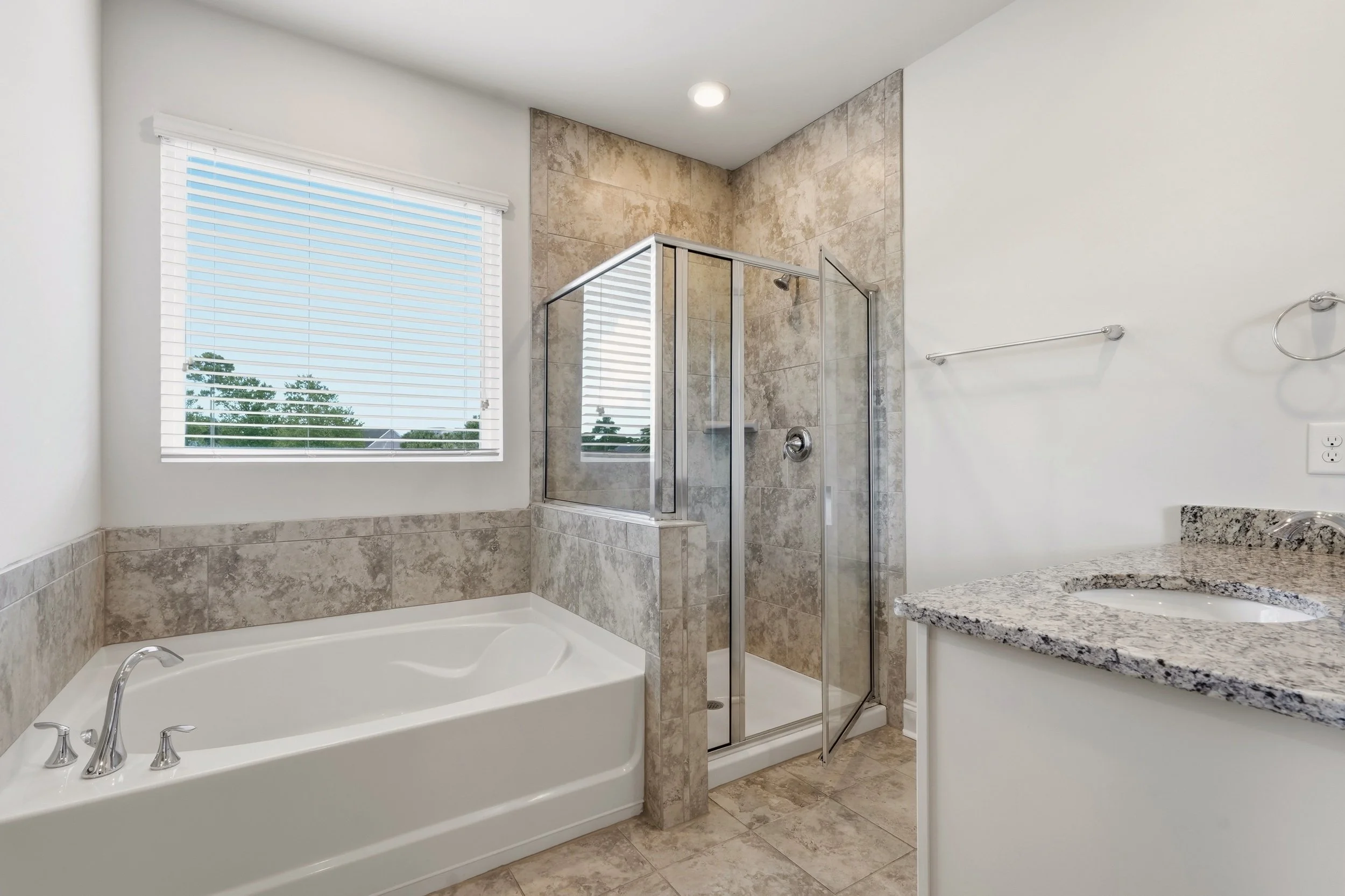 A modern bathroom featuring a bathtub, a corner glass shower with tiled walls, a granite countertop, and a window with white blinds overlooking trees.