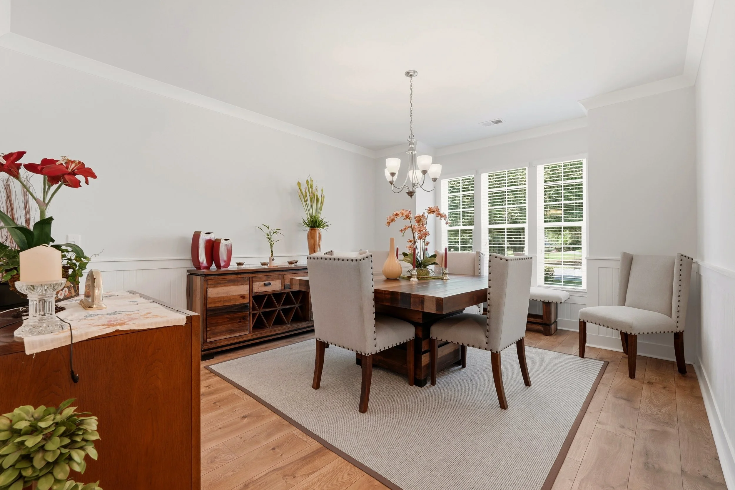 A dining room with white walls and a large window with white blinds. There is a wooden dining table surrounded by beige upholstered chairs with nailhead trim. On the table, there are vases with flowers, candles, and a decorative tray. A chandelier ha