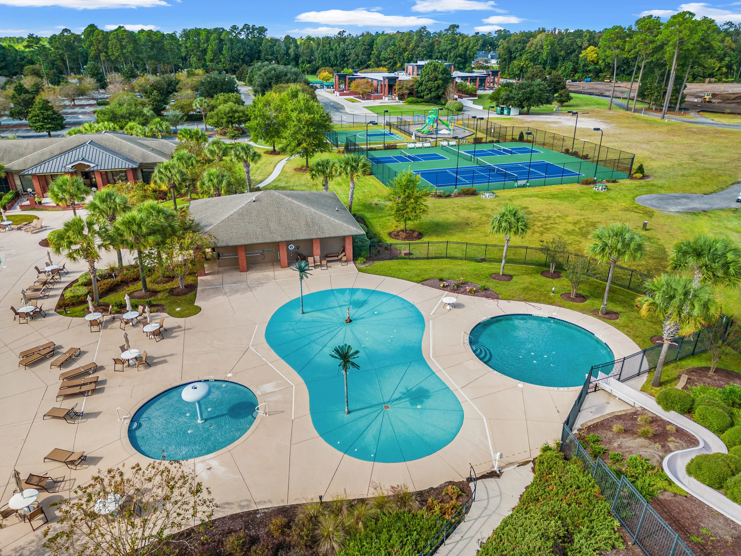 Aerial view of a recreational area with swimming pools, tennis courts, picnic tables, green trees, and surrounding buildings, under a partly cloudy sky.
