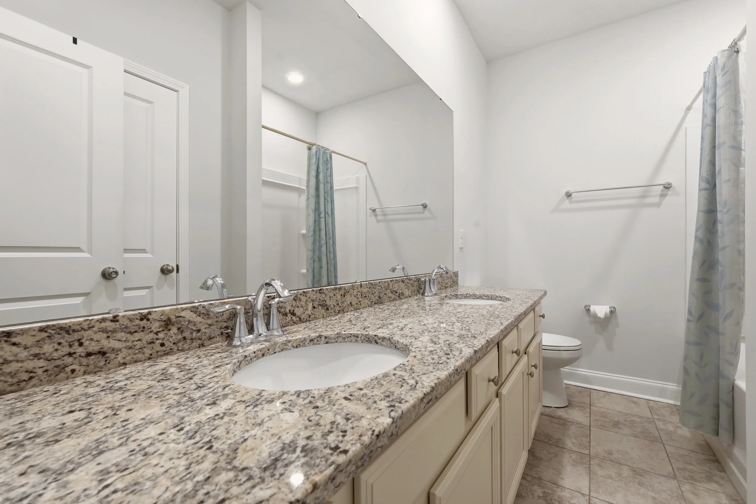 A bathroom with a granite countertop with dual sinks, a large mirror, white cabinetry, a shower with a curtain, a toilet, and white walls.