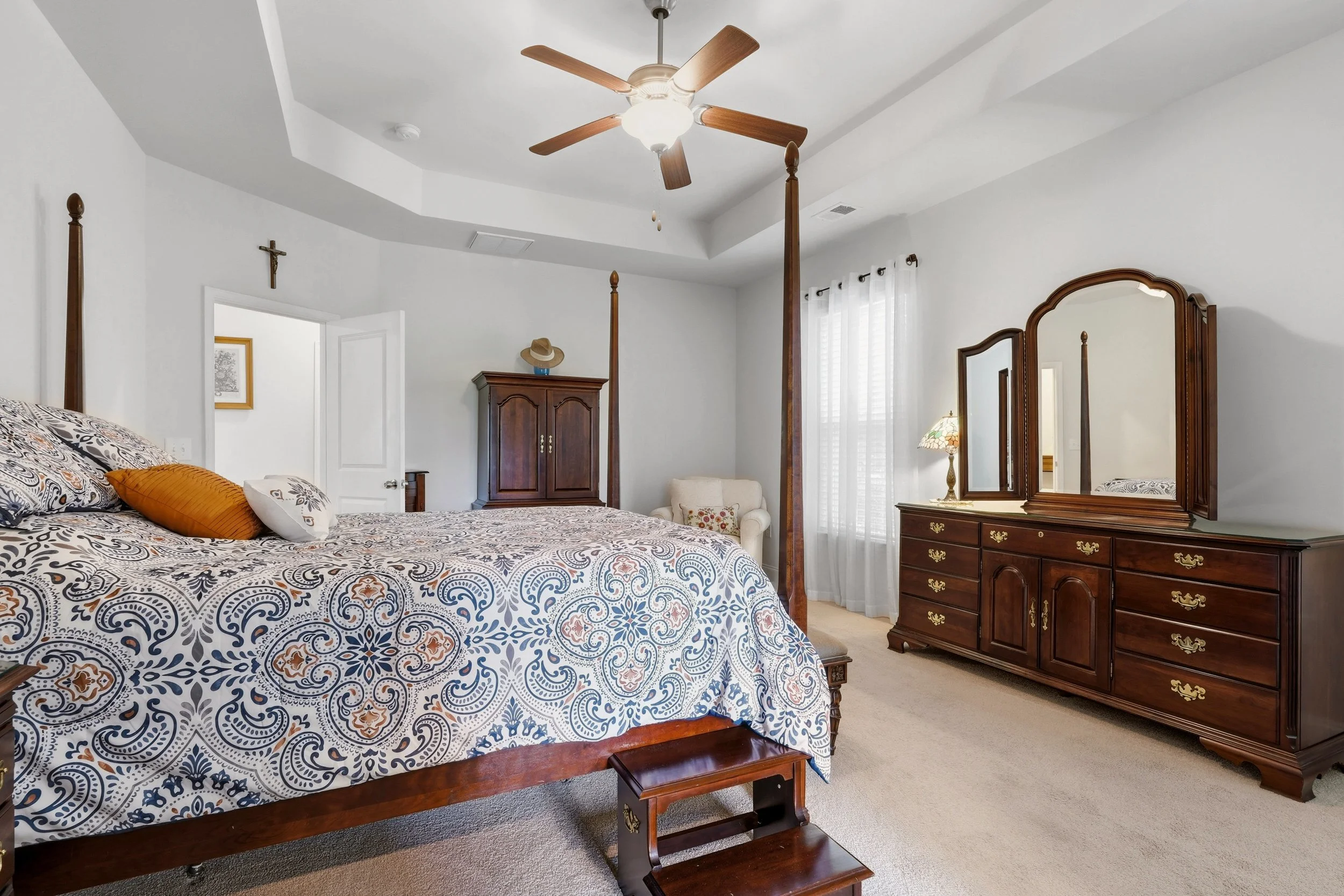 A bedroom with a wooden four-poster bed with patterned bedding and pillows. There is a wooden dresser with a mirror, a small chair, and a wooden step stool. The room has white walls, a ceiling fan, a crucifix on the wall, and windows with white curta