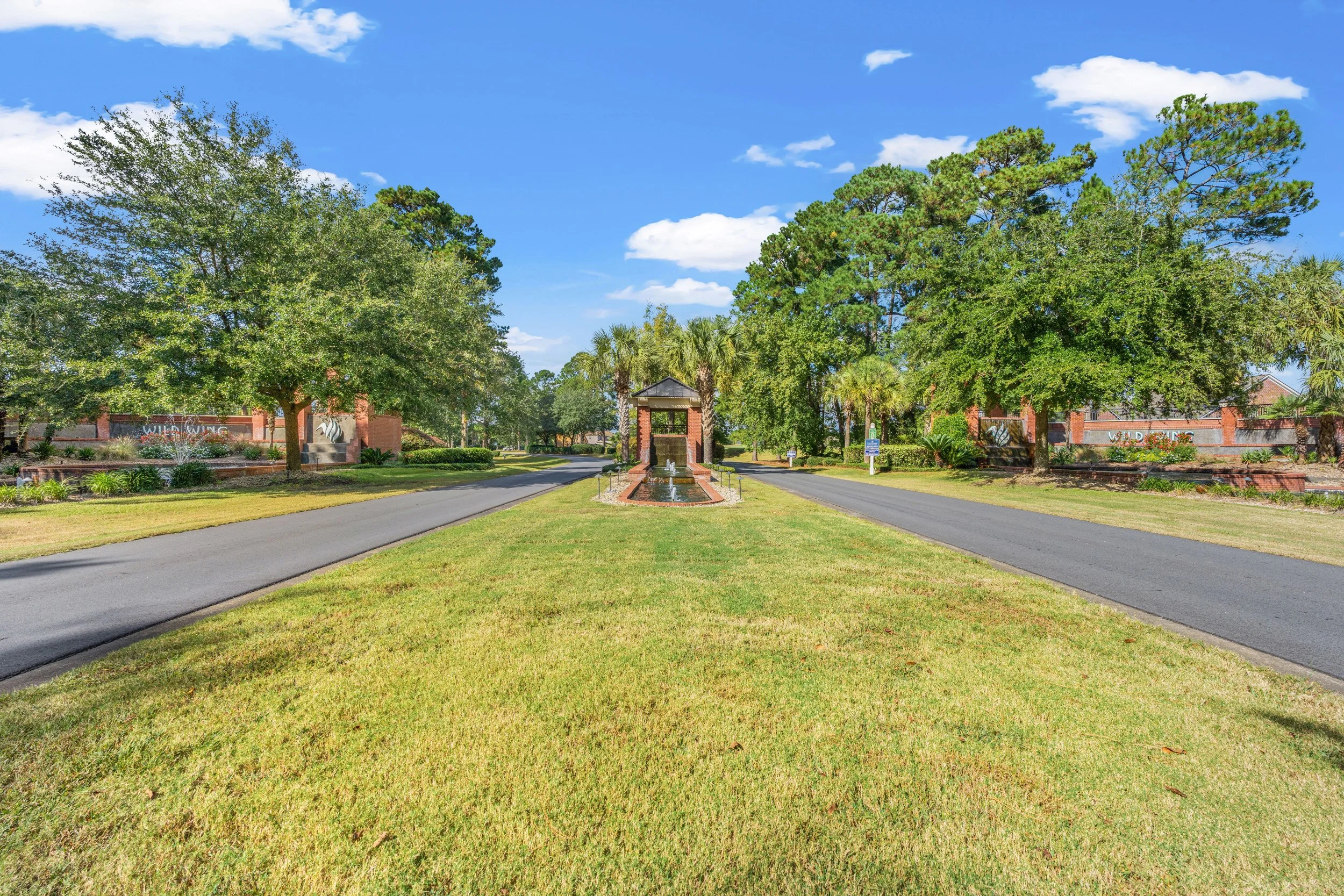 Entrance of a residential community with brick walls, lush green trees, a water fountain, and a paved driveway under a blue sky with scattered clouds.