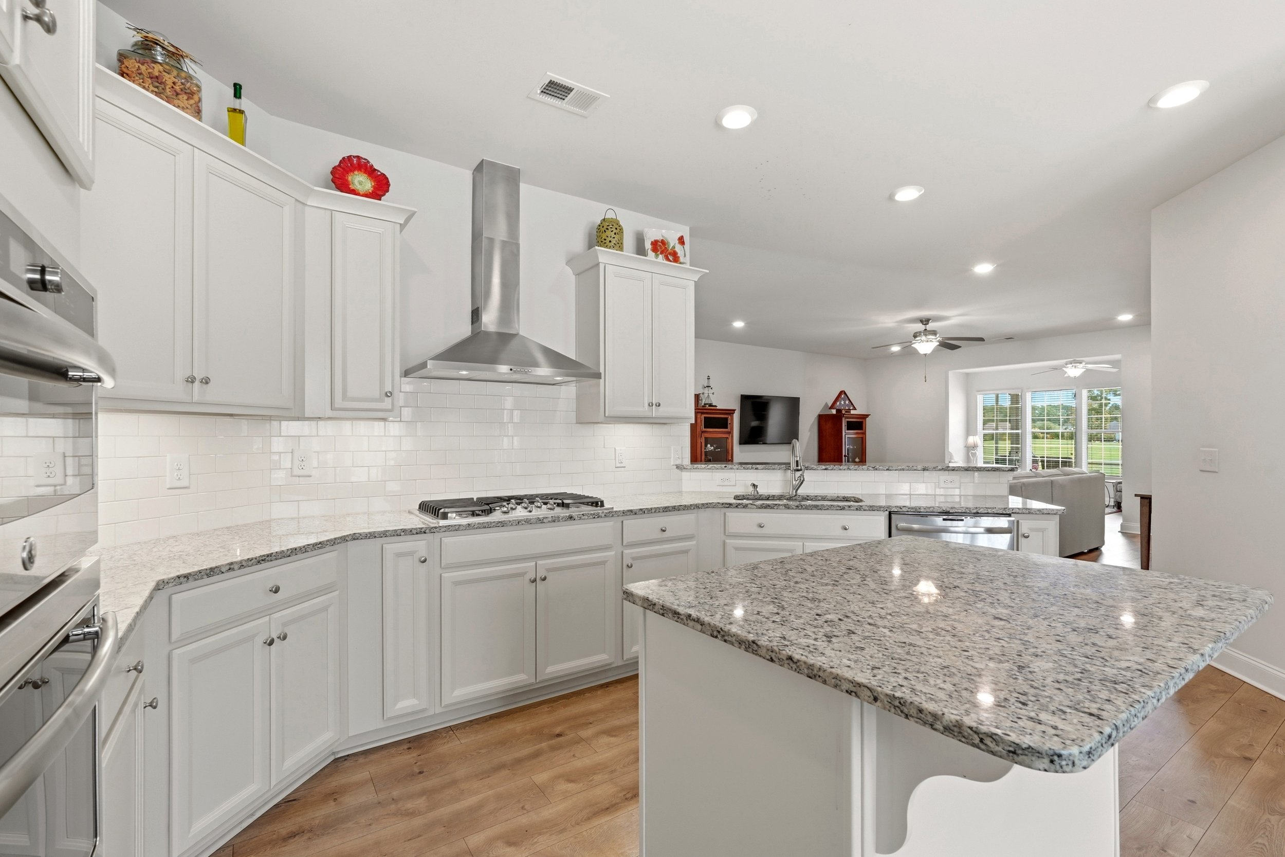 Modern white kitchen with granite countertops, white cabinetry, stainless steel appliances, a range hood, and a view into the living room with large windows and ceiling fans.