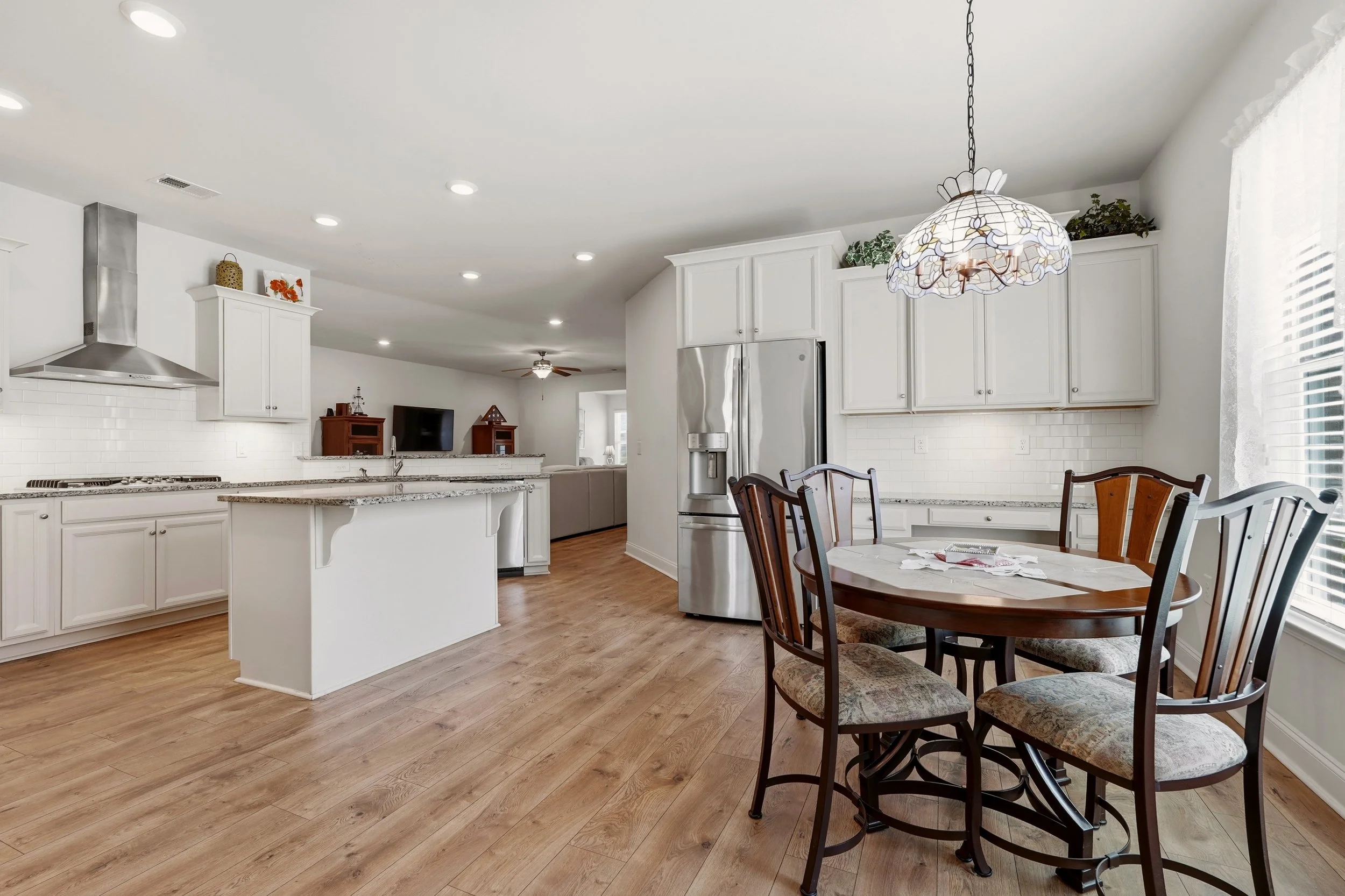 A kitchen and dining area with white cabinets, a stainless steel refrigerator, a marble countertop, a backsplash with white subway tiles, a hanging floral ceiling light, a round wooden table, six matching chairs, wood flooring, and a large window wit