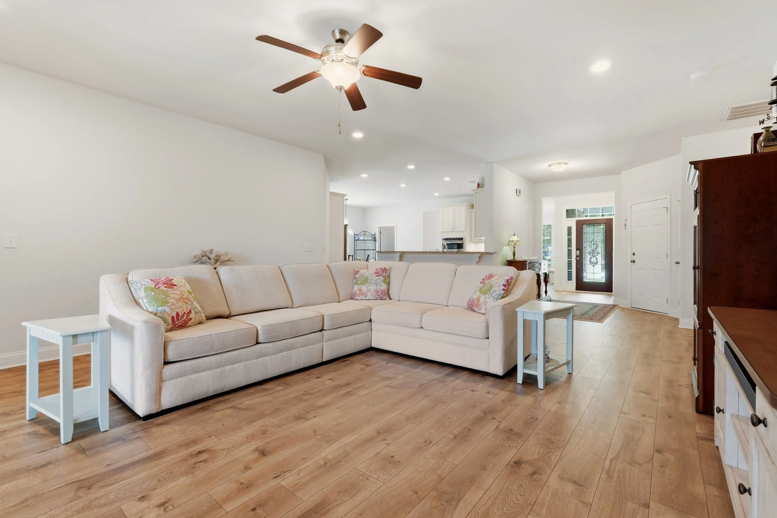 Living room with beige L-shaped sofa, white side tables, colorful floral pillows, wooden flooring, ceiling fan, and visible kitchen area in the background near the front door.