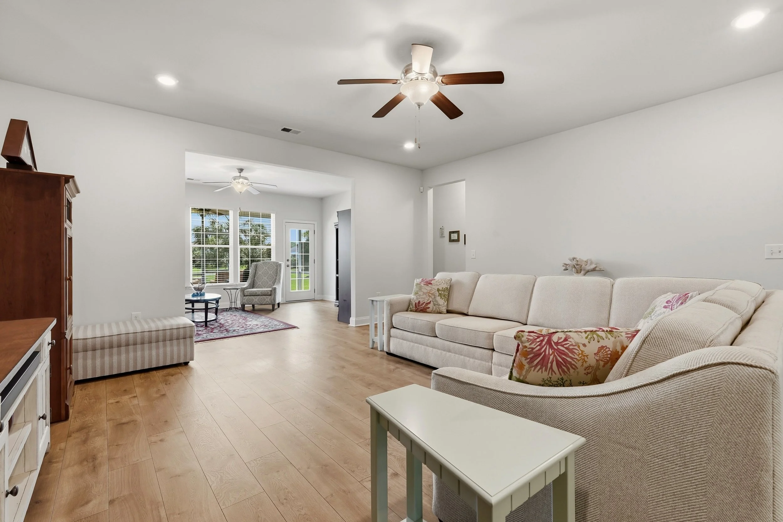 Living room with beige sofas, floral patterned pillows, wooden floor, ceiling fans, large windows, and a door leading outside.