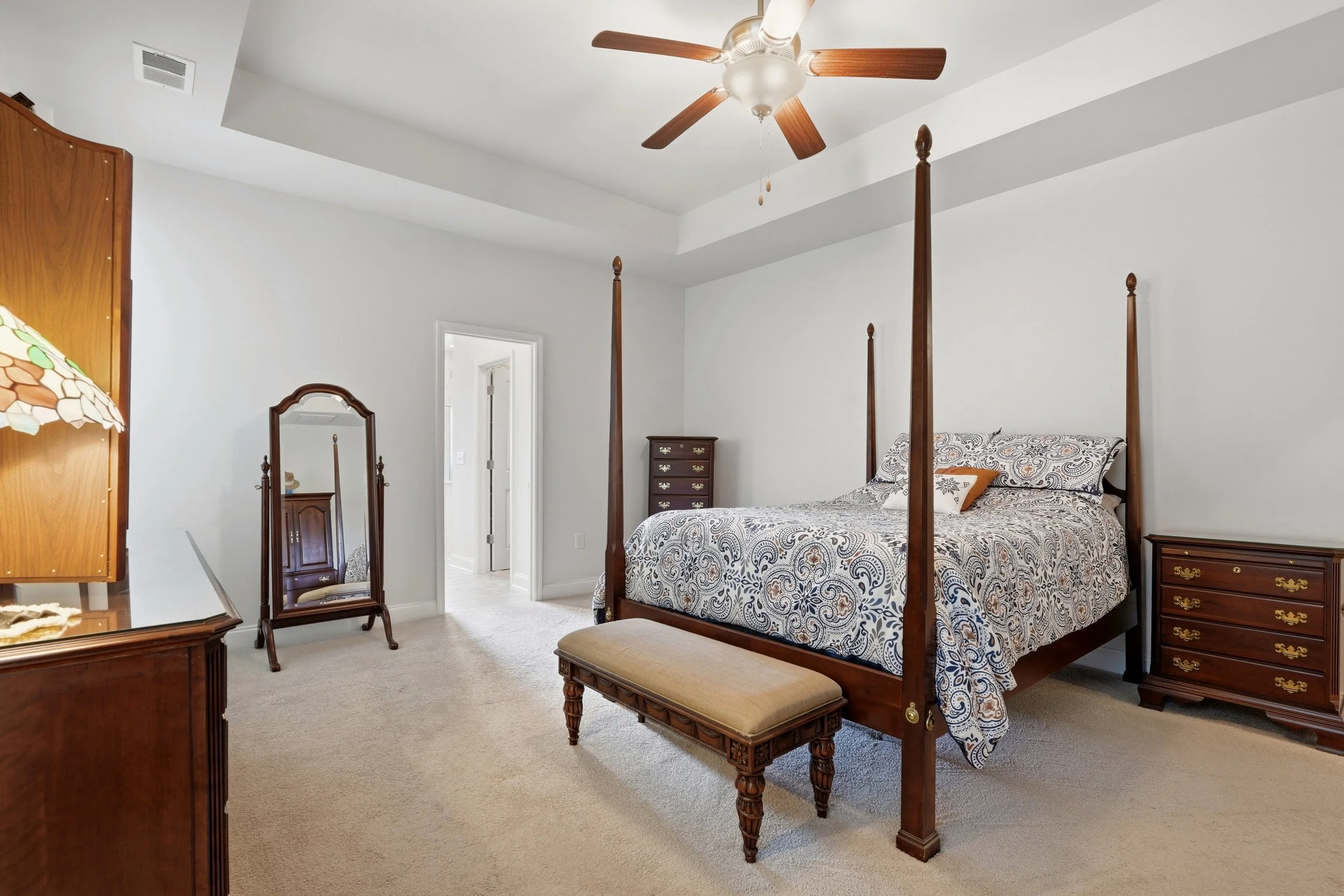 A bedroom with a four-poster wooden bed, a matching dresser and nightstands, a standing mirror, a bench at the foot of the bed, and a ceiling fan with wooden blades. The room has white walls, a beige carpet, and an open door leading to another room.