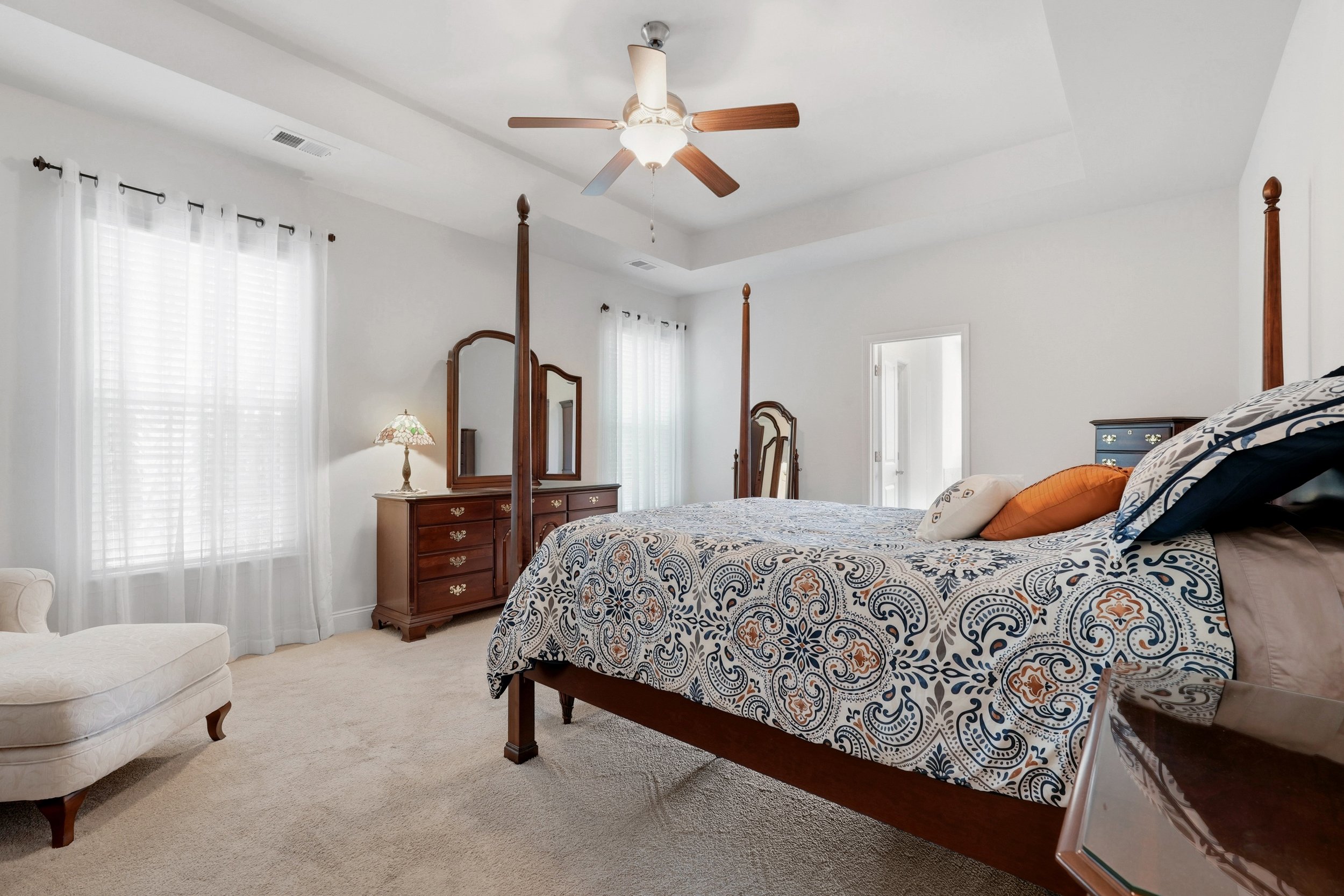 A bedroom with a four-post bed covered in a patterned quilt, a dresser with a mirror, a table lamp, and sheer curtains on the windows. There's a ceiling fan with lights and a beige armchair in the corner.