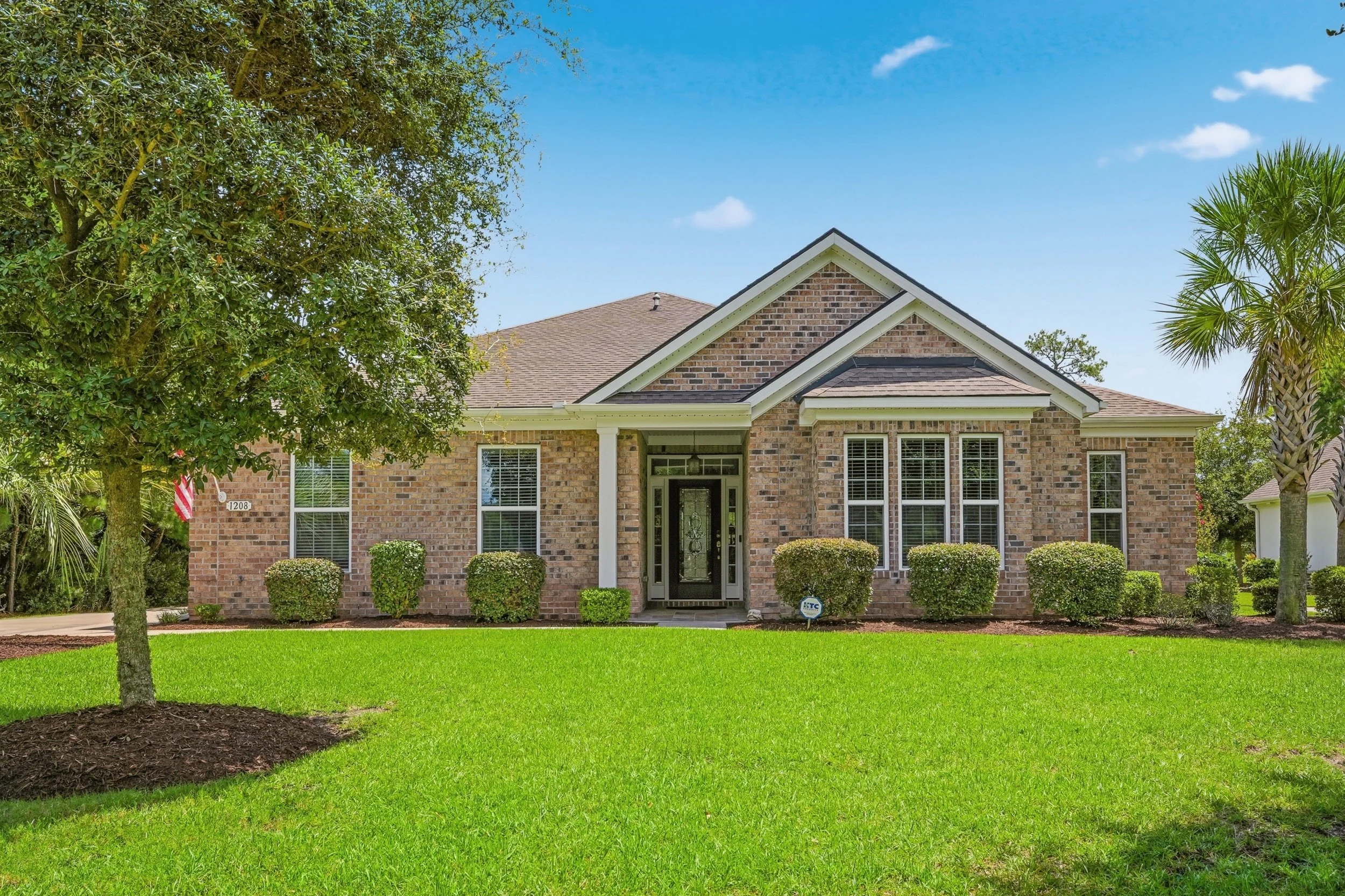 A brick house with a front entrance featuring a black door, white trim, and three windows with white shutters. The house has a sloped roof, and the yard has green grass, neatly trimmed bushes, and mature trees.