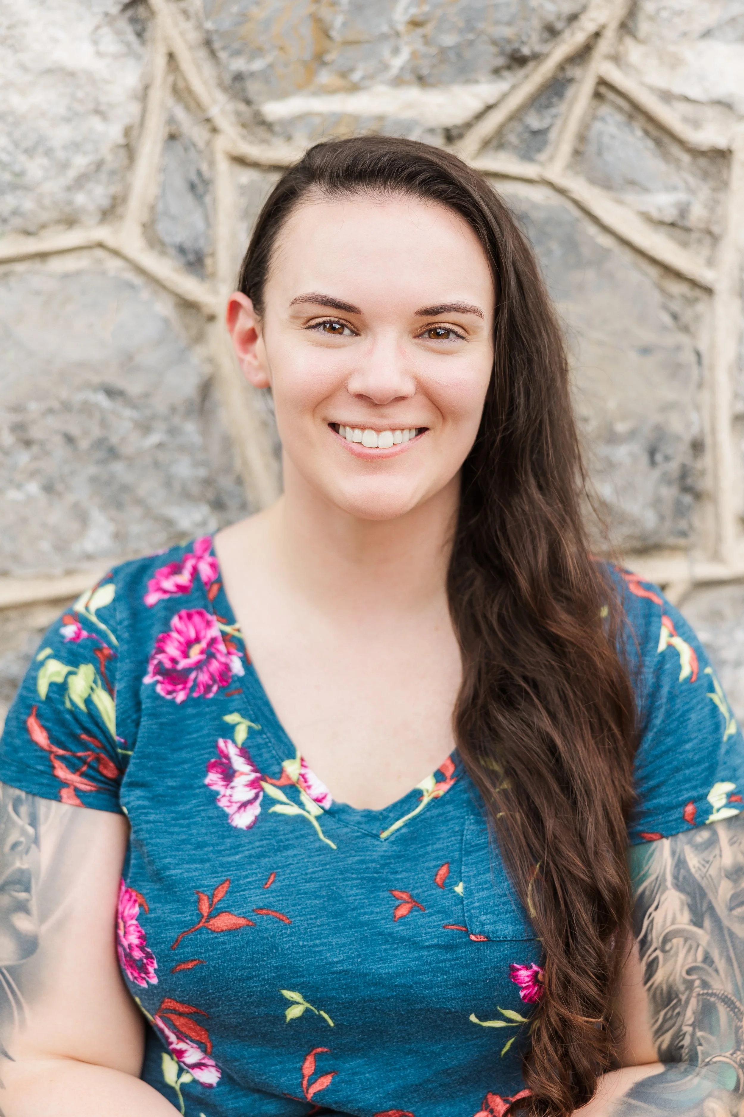A young woman with long brown hair smiling, wearing a blue floral shirt, standing against a stone wall.