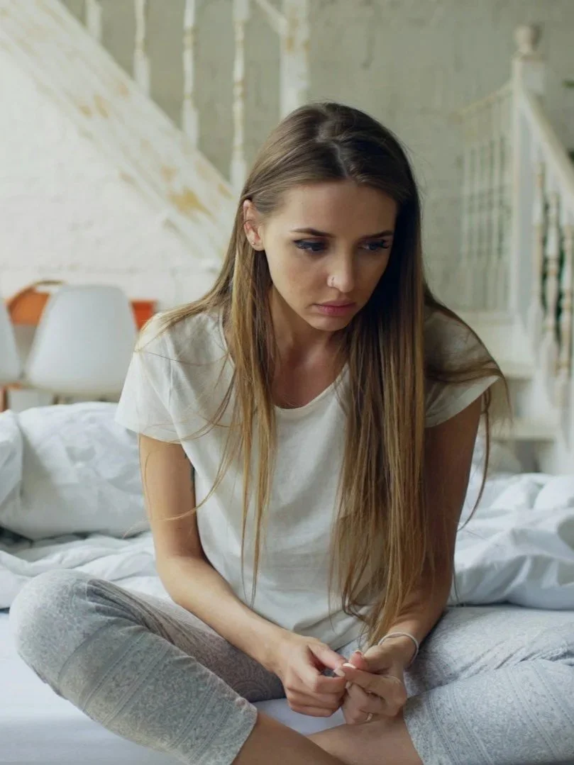 A woman with long brown hair sitting on a bed, looking down with a contemplative or worried expression, in a room with white walls and a staircase in the background.