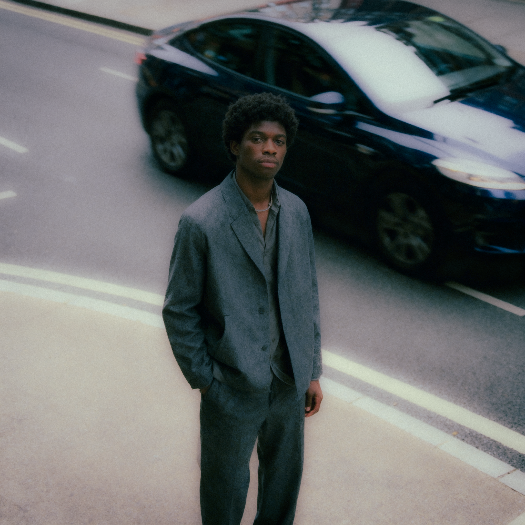 __Take inspiration from Tyler Mitchell photography. full body portrait of dark skinned male model in london next on the side of a road as a car passes behind him. Model styled in simple but effortlessly cool clothes, similar to Jill Sander .png