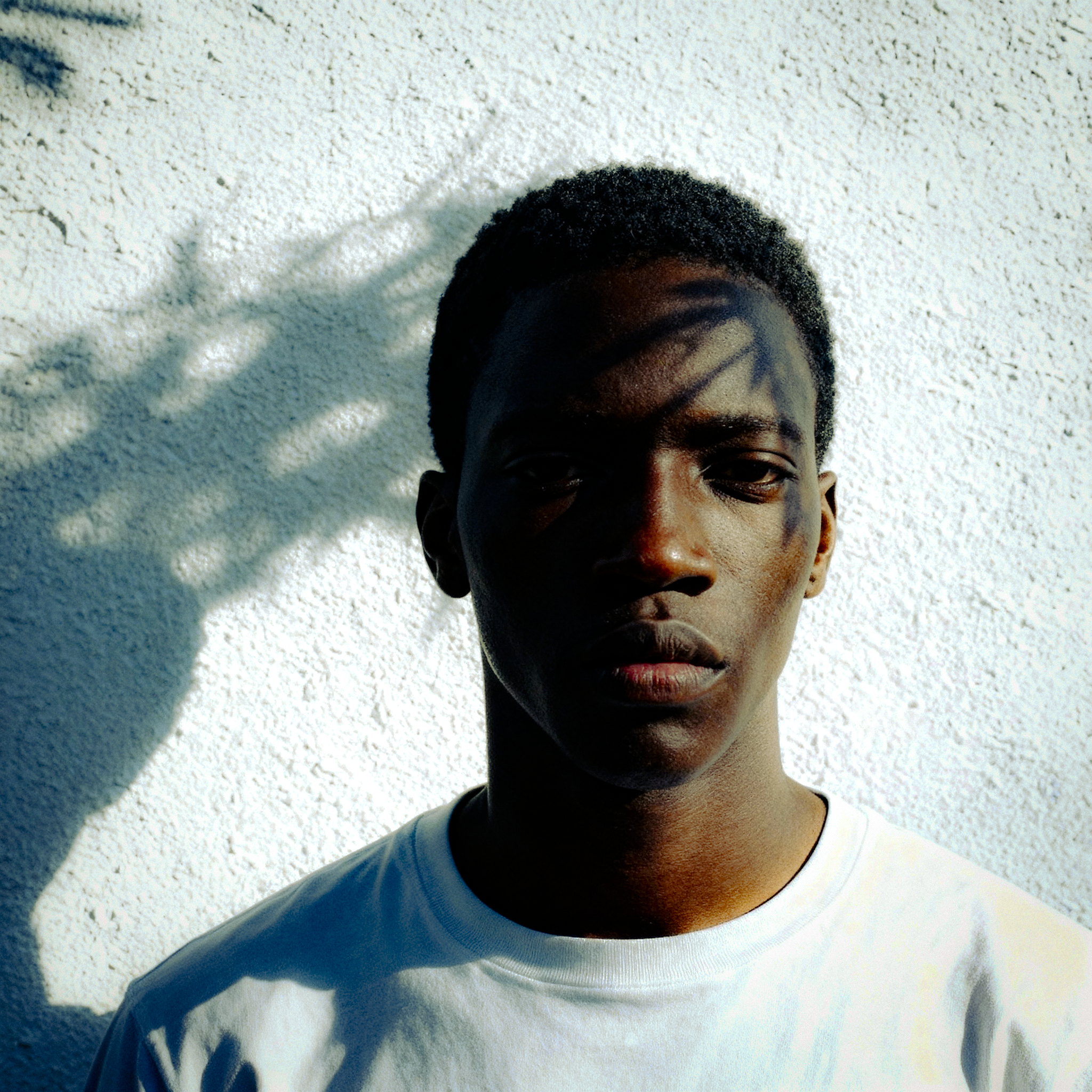 __Take inspiration from Tyler Mitchell photography. Close up portrait of dark skinned male model against a white textured wall. Model styled in simple but effortlessly cool shirt. Beautiful cool daylight grazing the models left side of chee.png