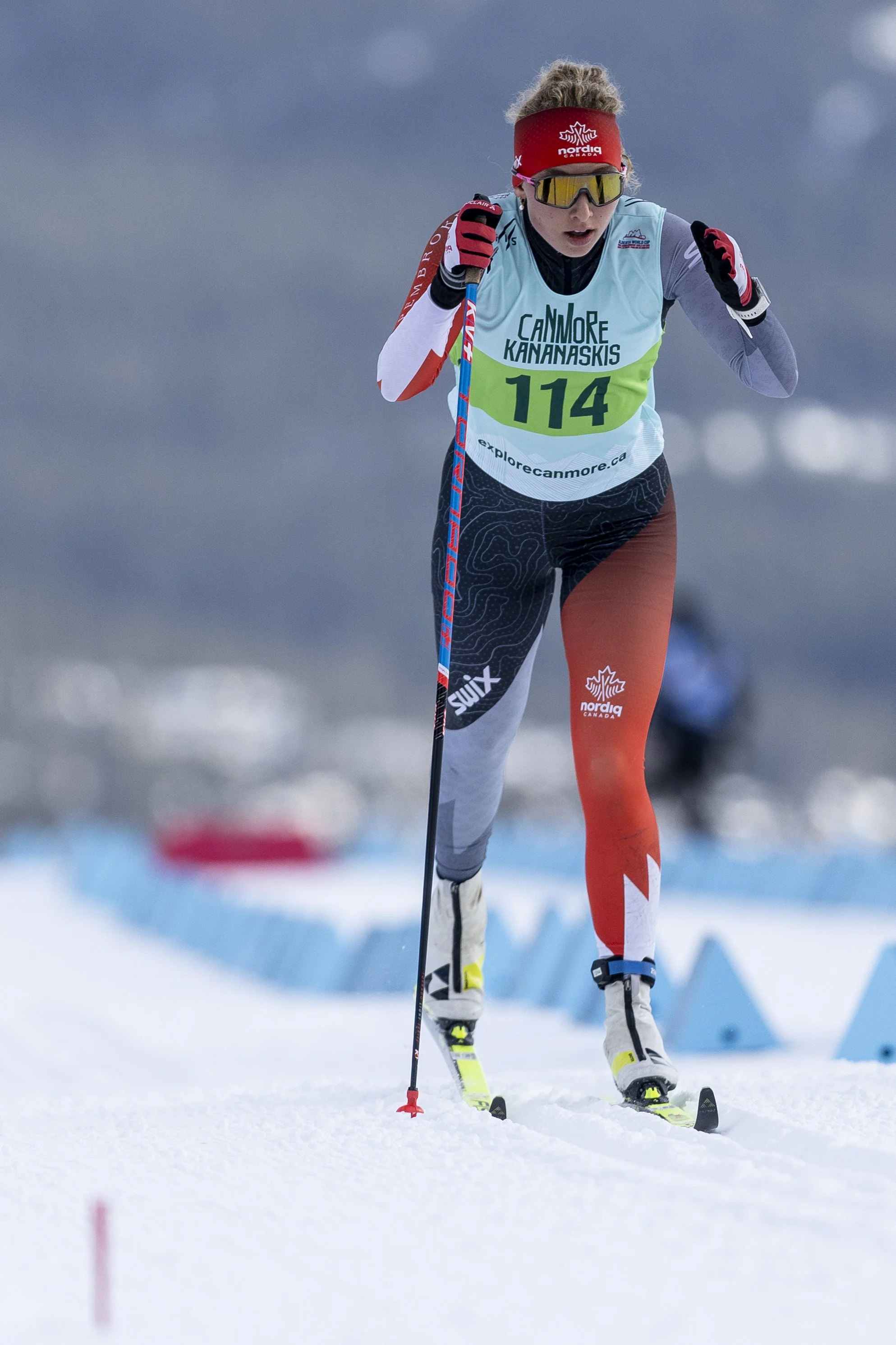 A female cross-country skier in racing gear, wearing sunglasses, a red headband, and a race bib number 114, skiing on snow during a race in cloudy weather.