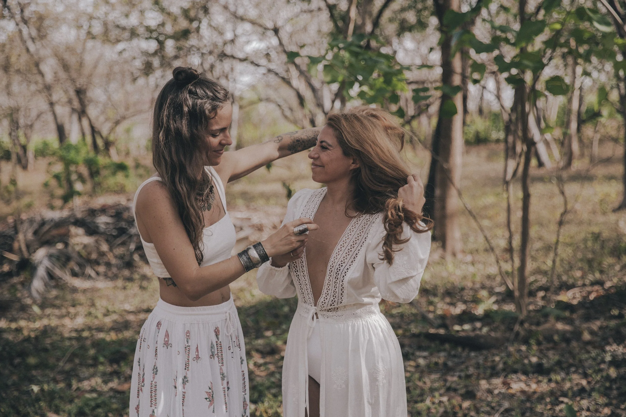 Two women with long hair, one with brown hair and the other with dark hair, stand close together outdoors in a wooded area. They are facing each other, smiling softly, and seem to be sharing a meaningful moment. The woman with dark hair is playing with the blonde woman's hair.