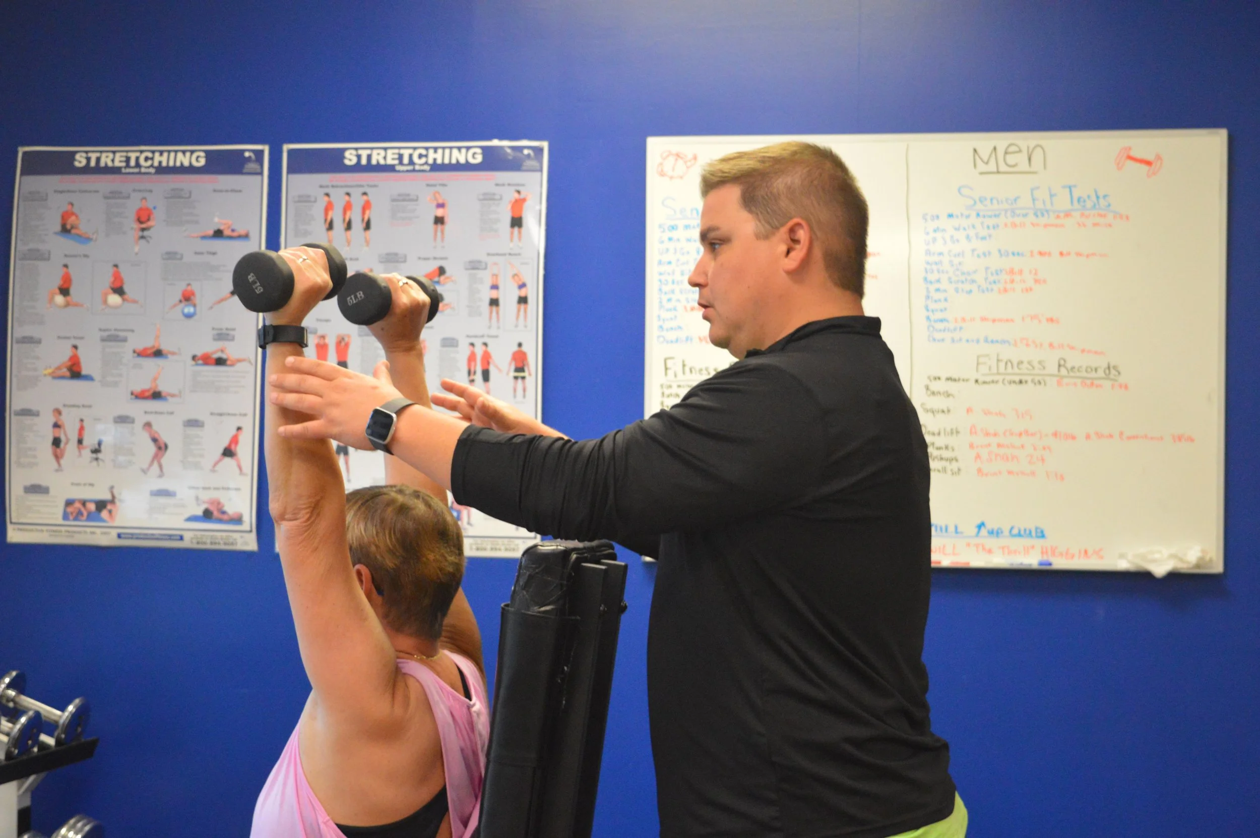 A male fitness instructor assists an older woman with an overhead dumbbell exercise in a gym, with fitness charts on the blue wall behind them.