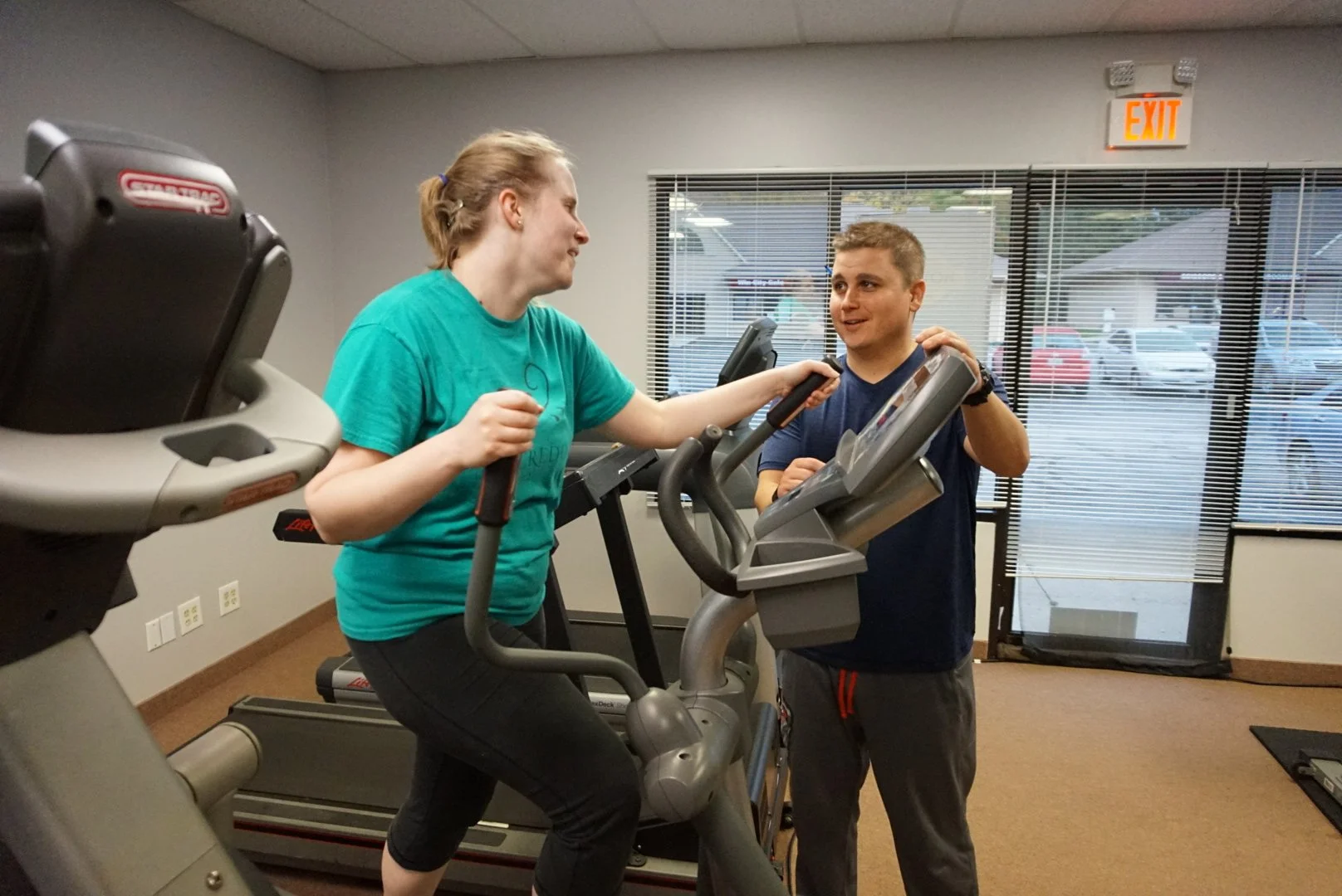 A woman on a treadmill with a man beside her, helping or encouraging her, in a gym or fitness center.
