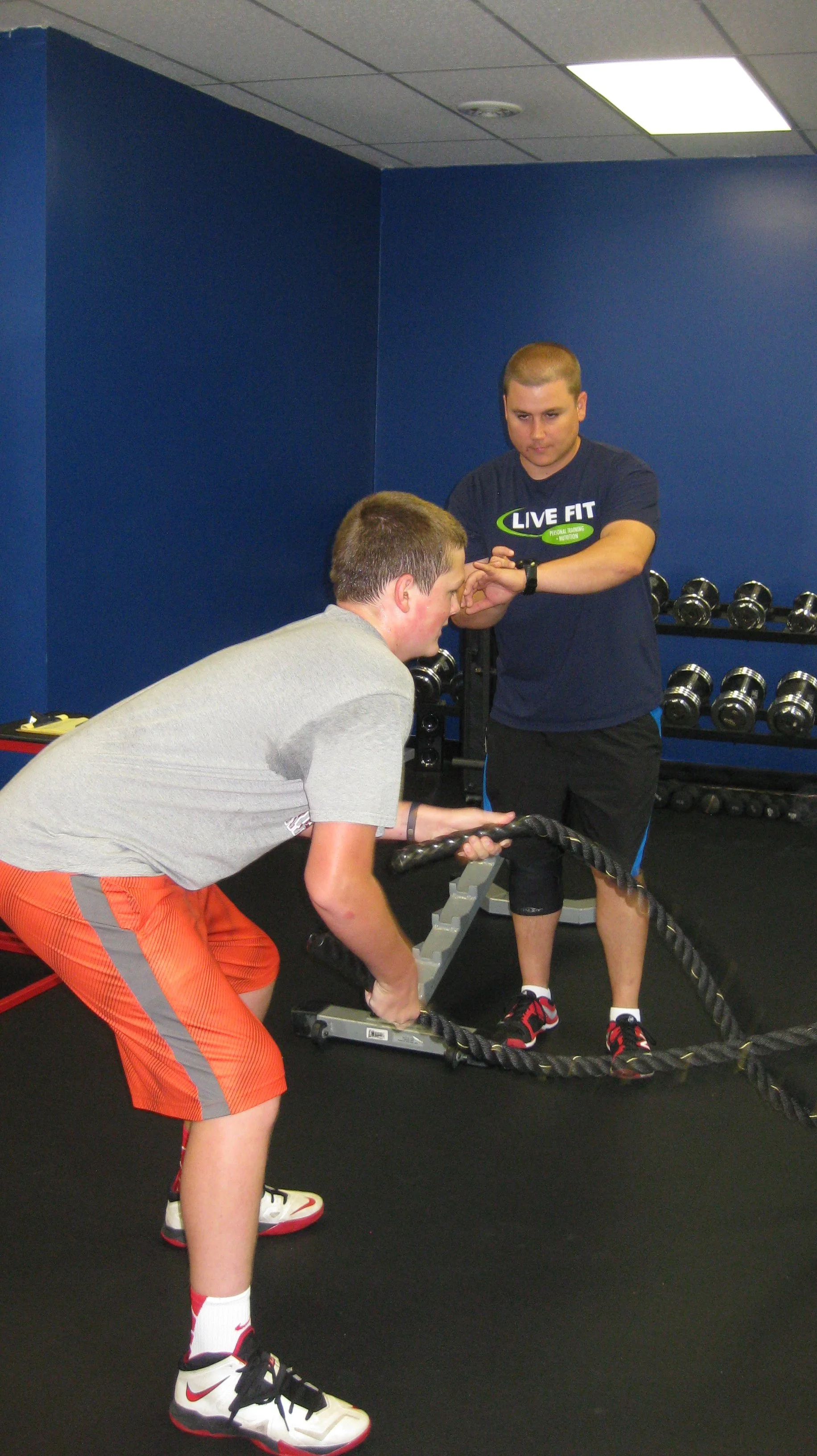 A male trainer guides a teenage boy through a workout using battle ropes at a gym with blue walls and a rack of dumbbells.