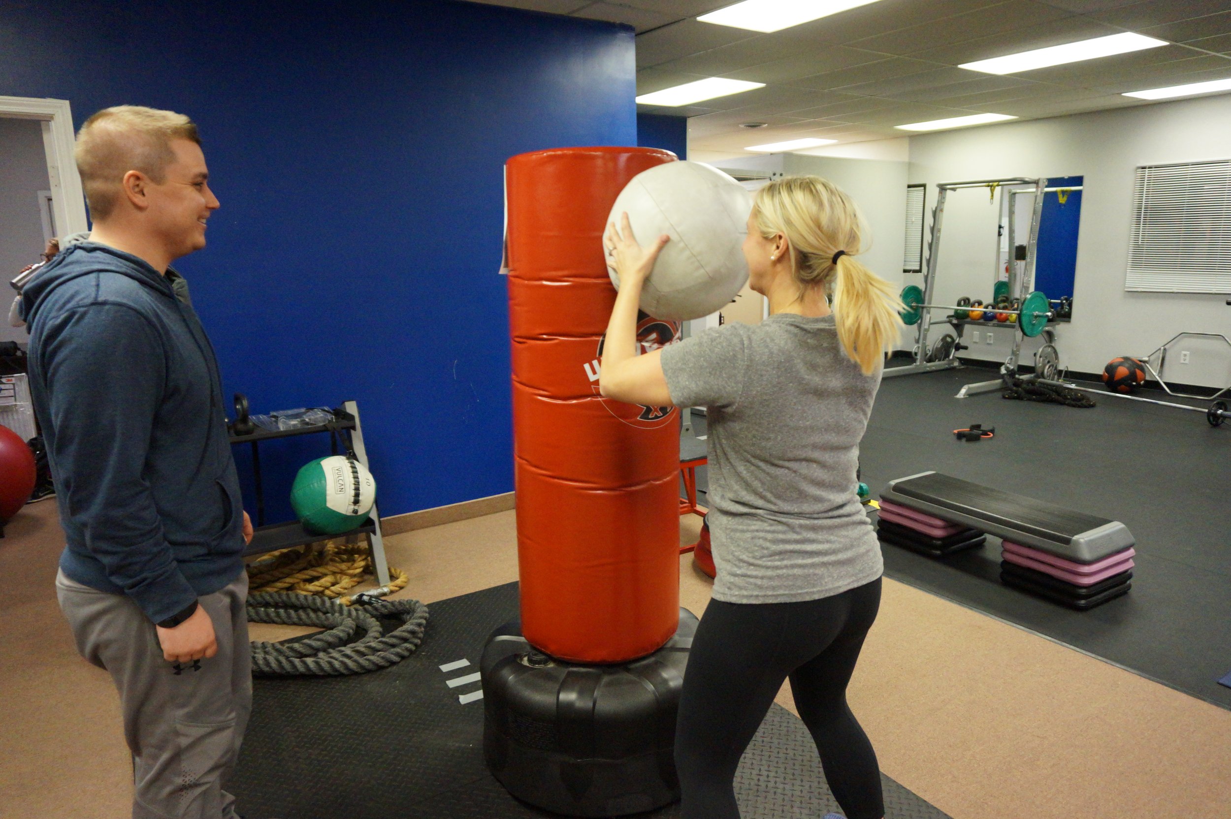 A woman is hitting a punching bag with a medicine ball while her trainer watches and smiles in a gym.