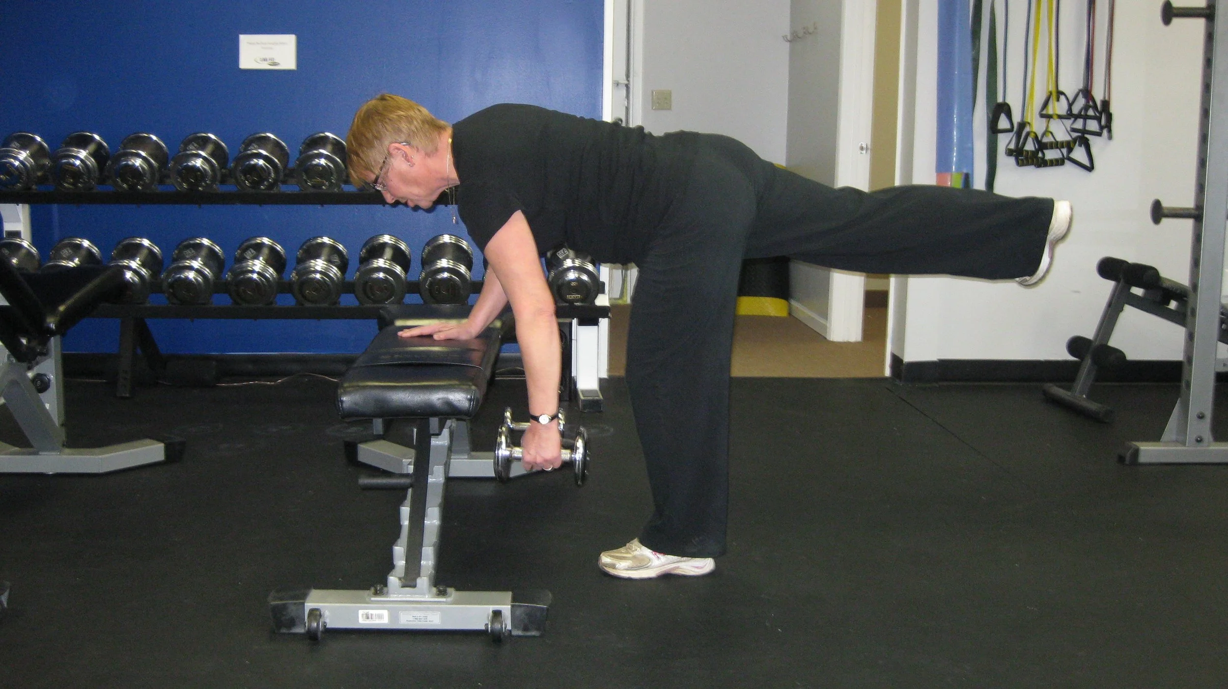 A woman exercising in a private personal training studio, using a dumbbell while supporting herself with one hand on a bench and the opposite leg extended straight back.