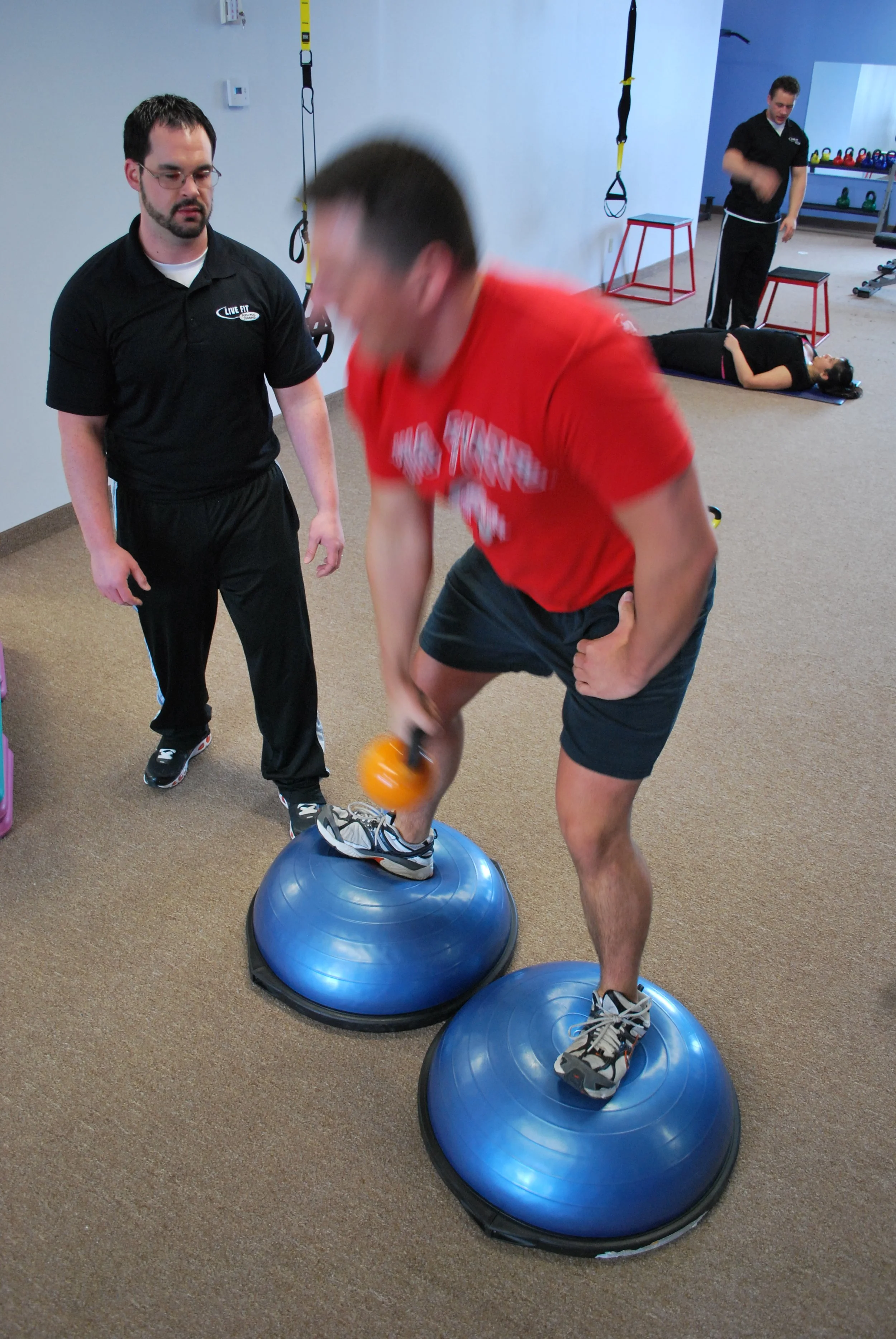 A man in red shirt and black shorts balancing on two half-sphere balance trainers while holding a small orange kettlebell in a fitness training room. A trainer in black outfit stands nearby, observing. In the background, a woman is exercising on a ma