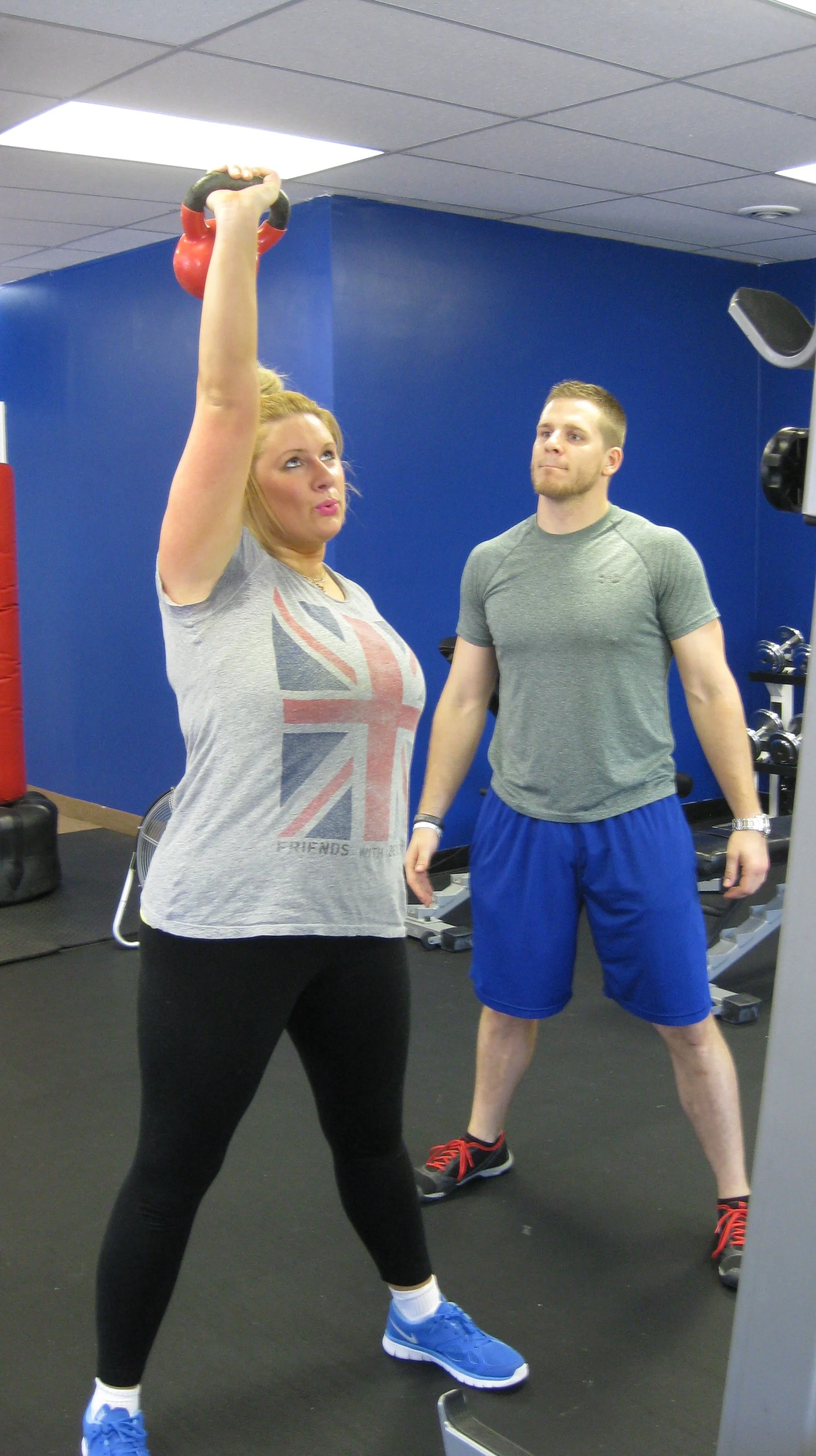 A woman performing an overhead kettlebell exercise with a trainer watching.