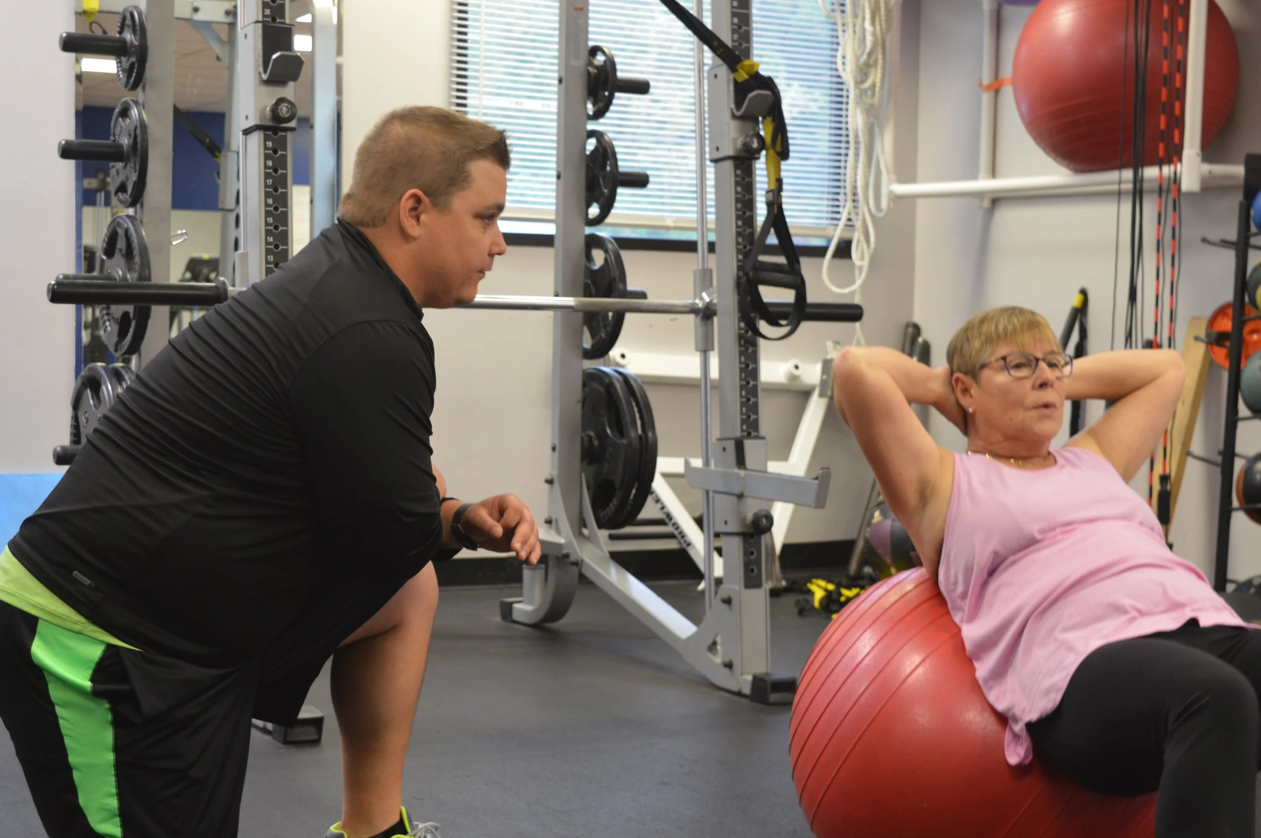 A woman in a pink shirt is doing crunches on an exercise ball while a trainer in black fitness attire observes in a training studio with weights and fitness equipment in the background.
