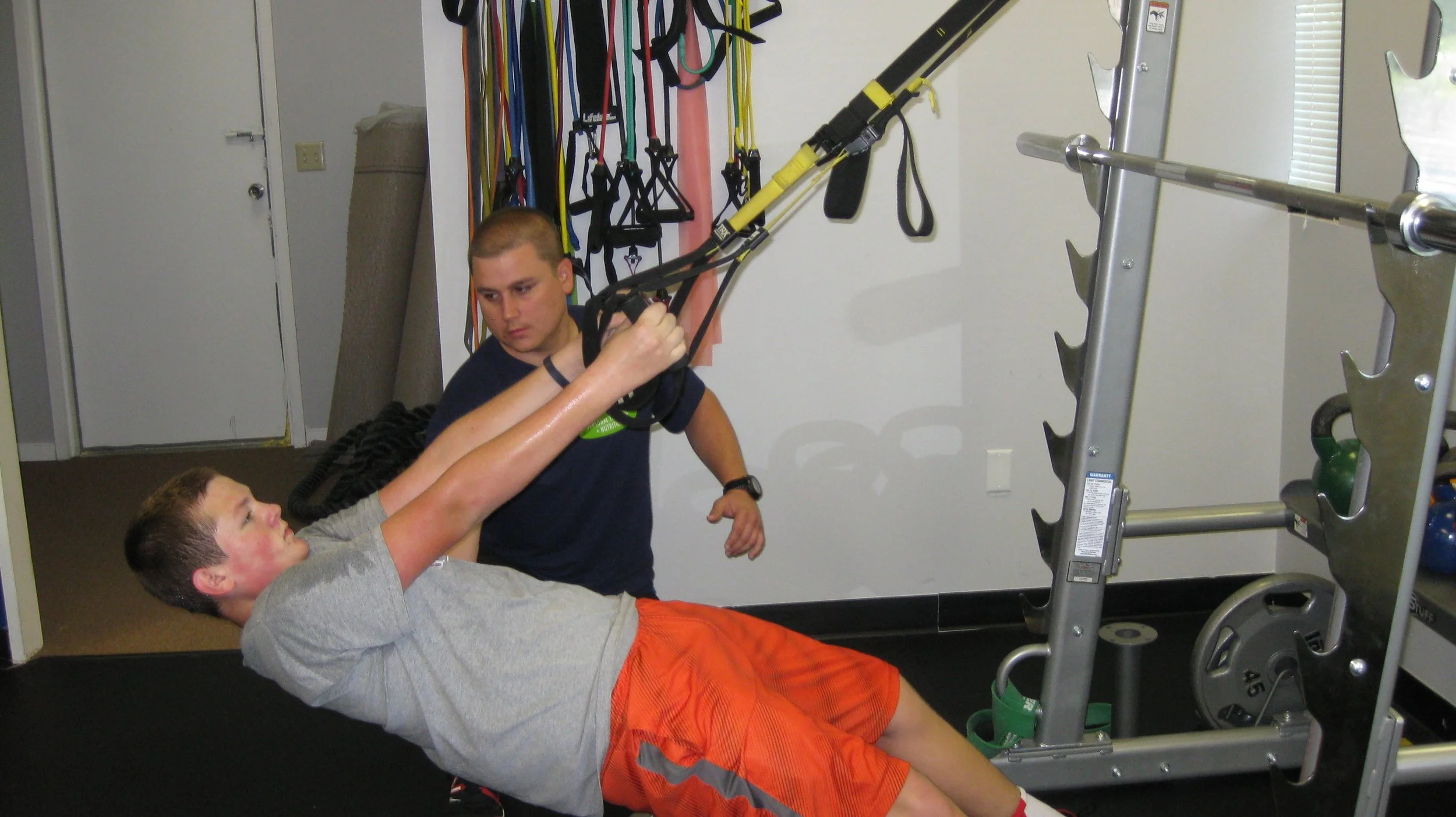 A man lying on his side performing a suspension exercise with assistance from his trainer in a gym. The fitness studio has resistance bands hanging on the wall and weightlifting equipment in the background.