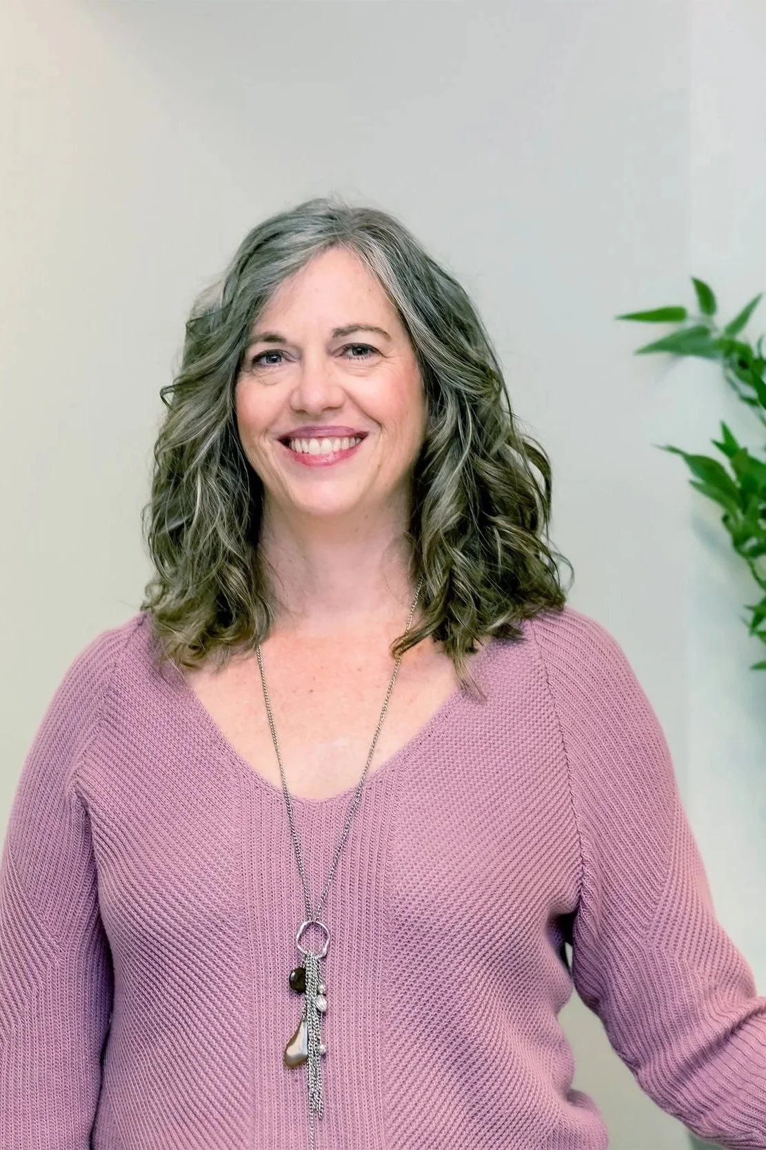 A woman with curly gray hair and a pink sweater, smiling for the camera, standing in front of a plain wall with a green plant on the right.