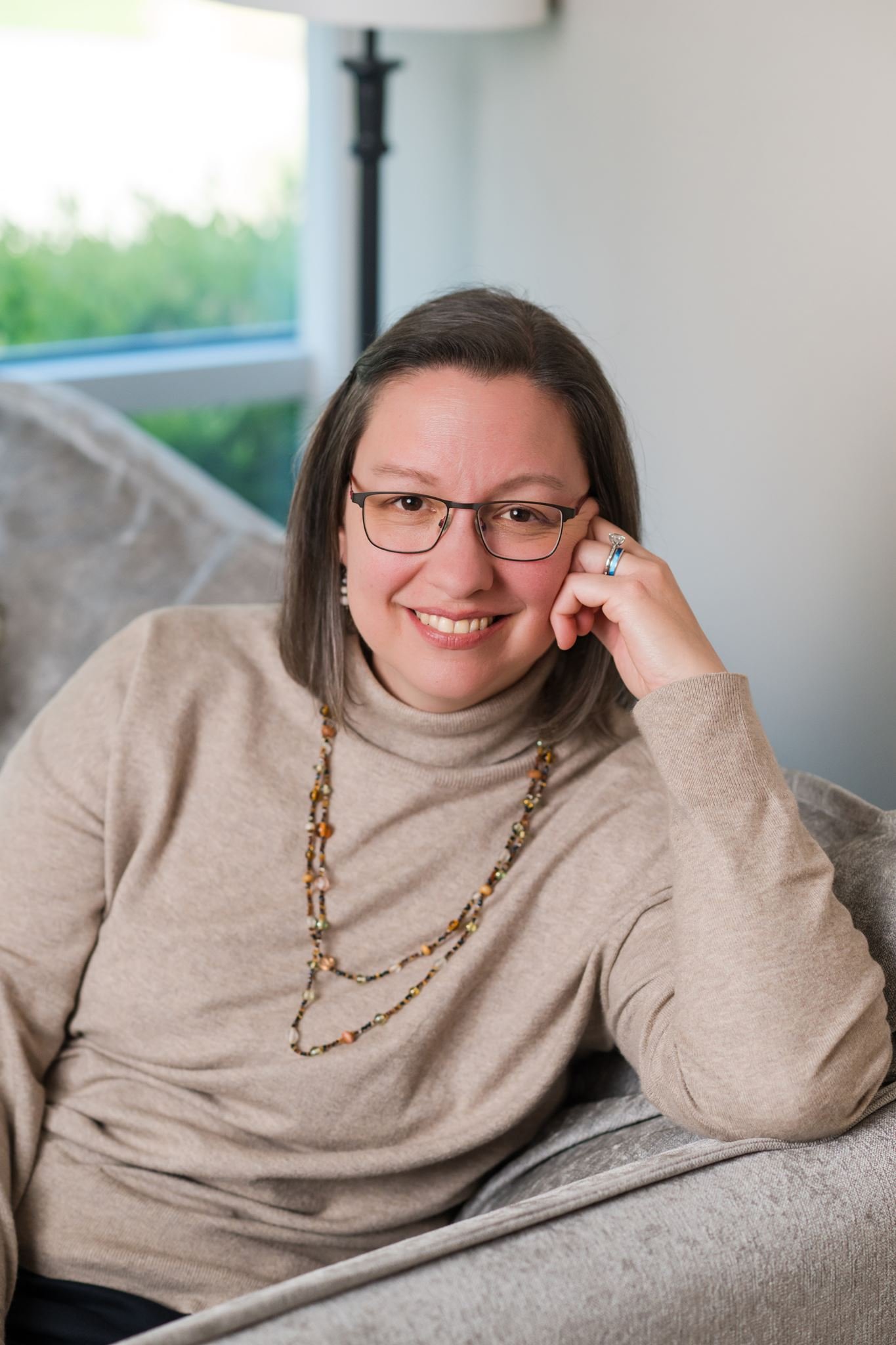 A woman with glasses and earrings, wearing a white top and a pink shawl, smiling in front of a white background.