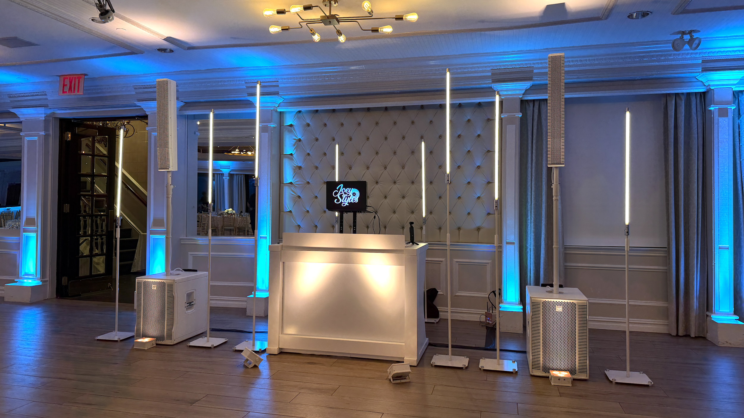 Event space with DJ booth and lighting setup, featuring a white DJ booth, vertical LED lights, two speakers on stands, and a small screen displaying 'Joey Style' amidst blue uplighting on walls.