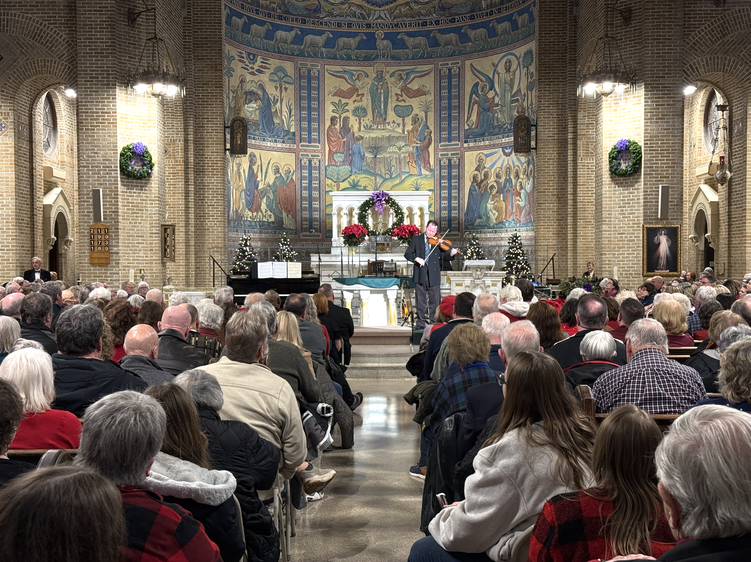 A concert inside a church with a large audience, a violinist performing on stage, decorated with Christmas wreaths, trees, and poinsettias, with a mural on the back wall depicting religious figures.