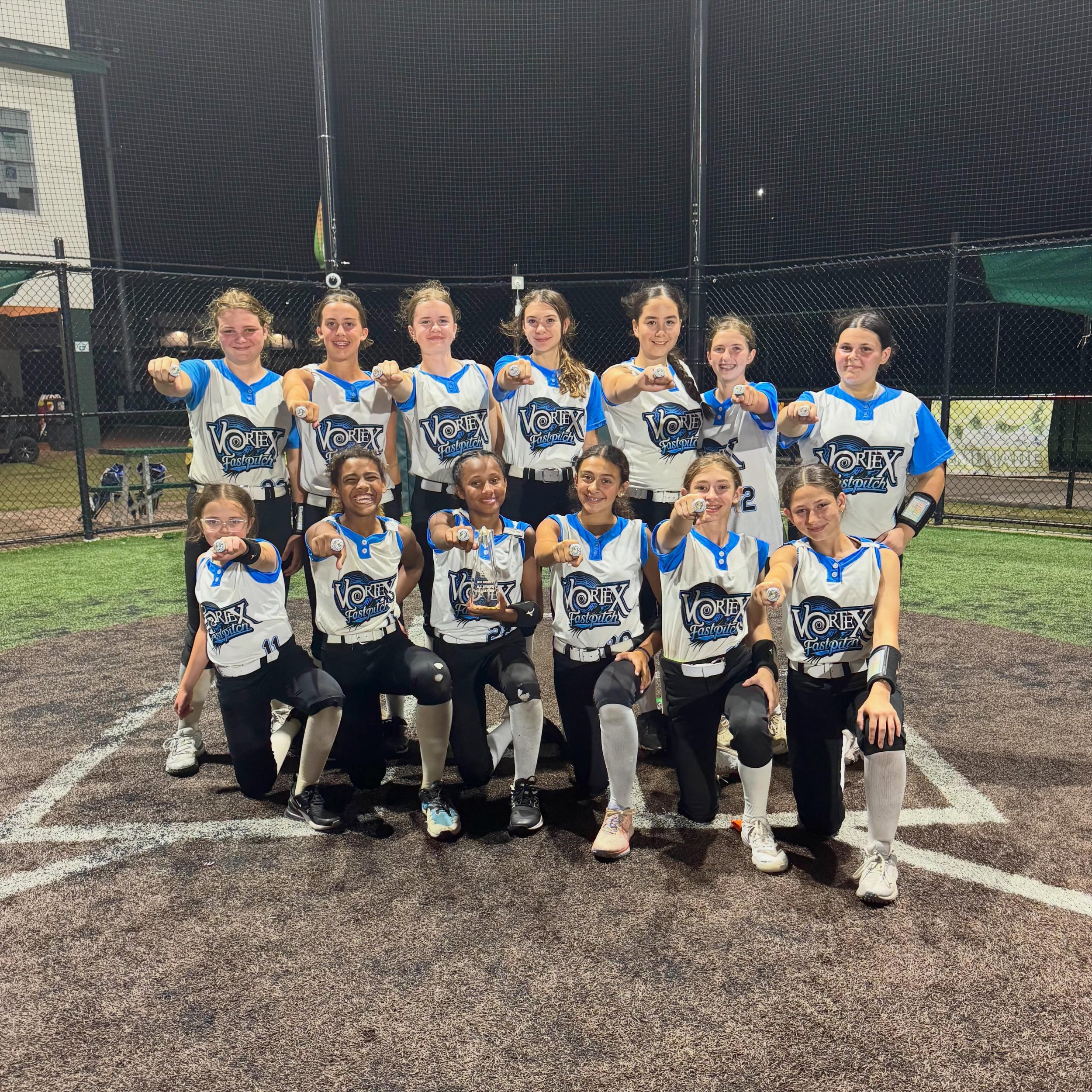 Young girls in colorful softball uniforms posing together on a field with a chain-link fence and trees in the background.