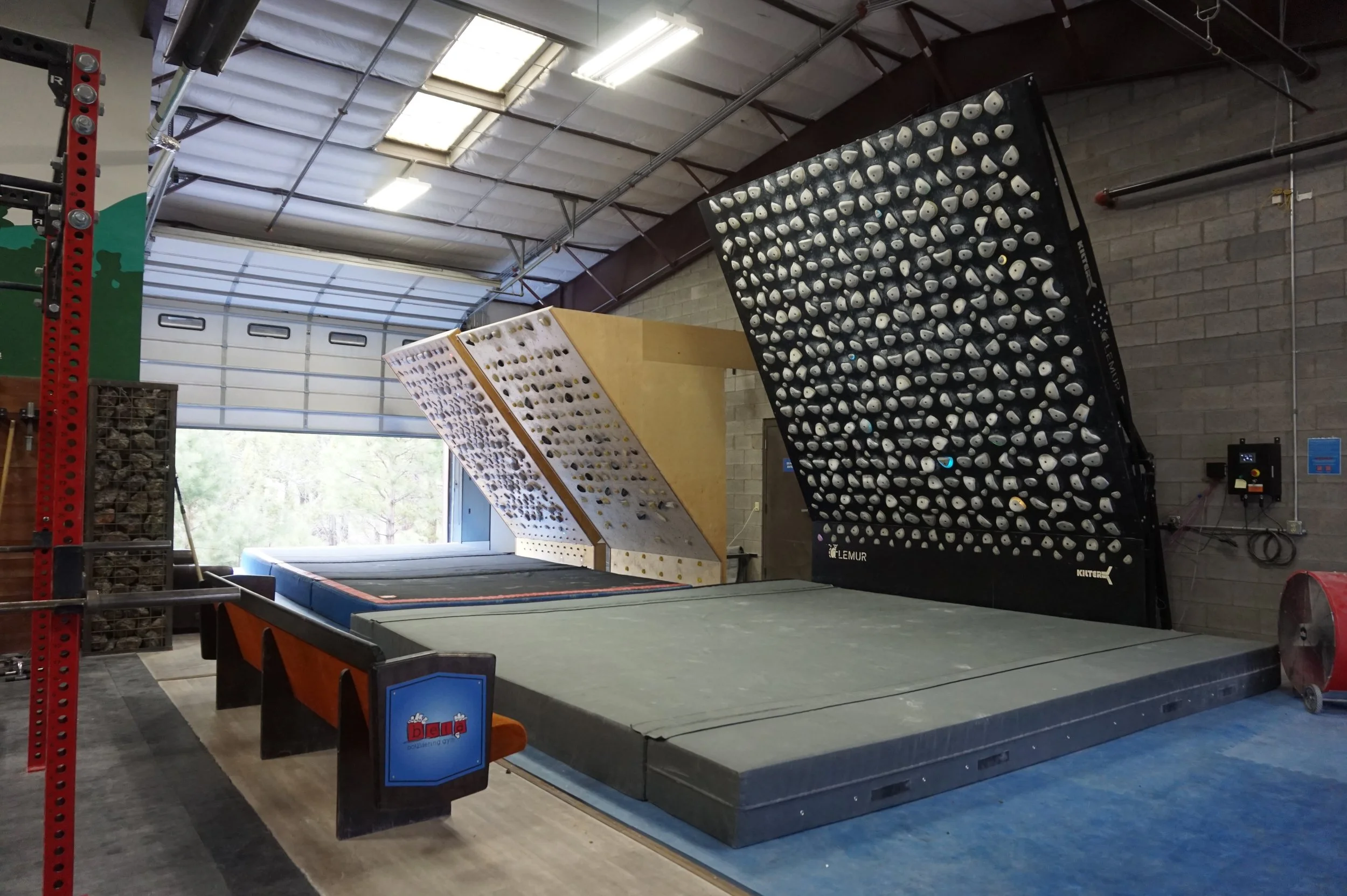 Indoor climbing gym with a large black climbing wall featuring numerous white holds, a smaller angled climbing wall with yellow holds, and a padded floor area for safety.