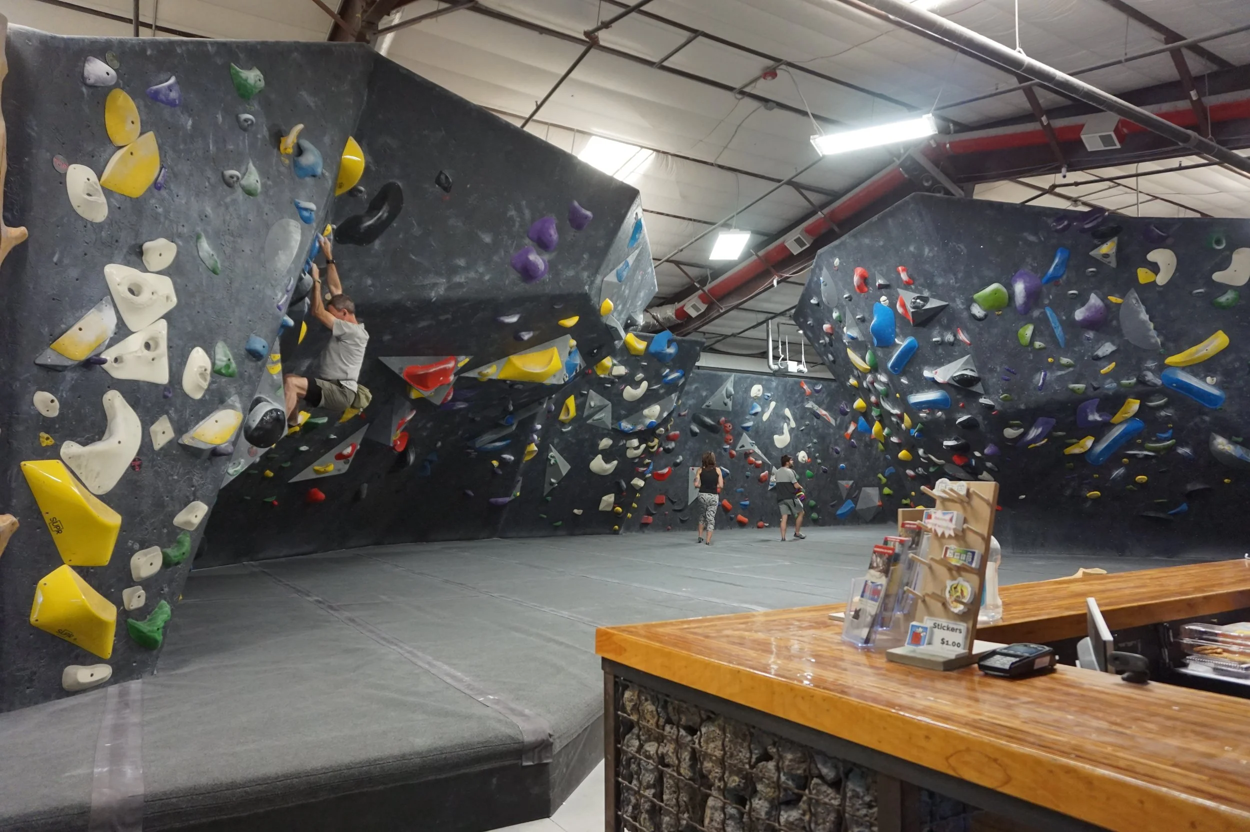 Indoor rock climbing gym with a man climbing on a gray climbing wall covered in colorful holds, and a table with stickers in the foreground.